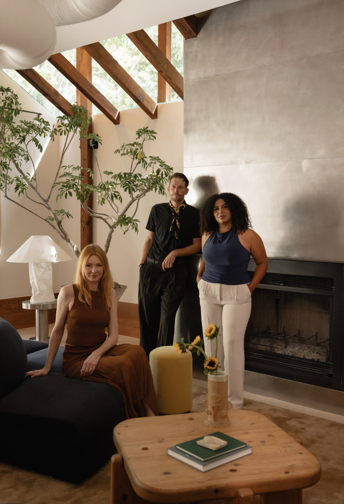 Four people are in a living room, three of them are standing and one sitting on a black couch. The room has a wooden table with a book and a small decorative item, a yellow ottoman with sunflowers, a lamp, and a tree. The background includes large windows, wooden beams, and a fireplace.