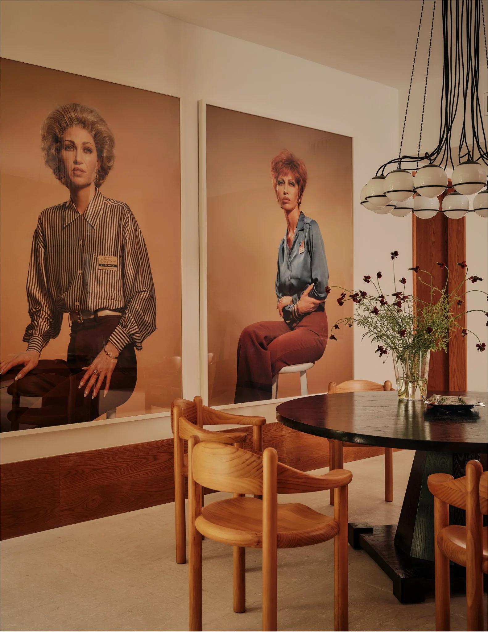Interior of a room with two large portraits of women on the wall, a modern chandelier hanging from the ceiling, a glass vase with dark flowers on a round black table, and wooden chairs around the table.