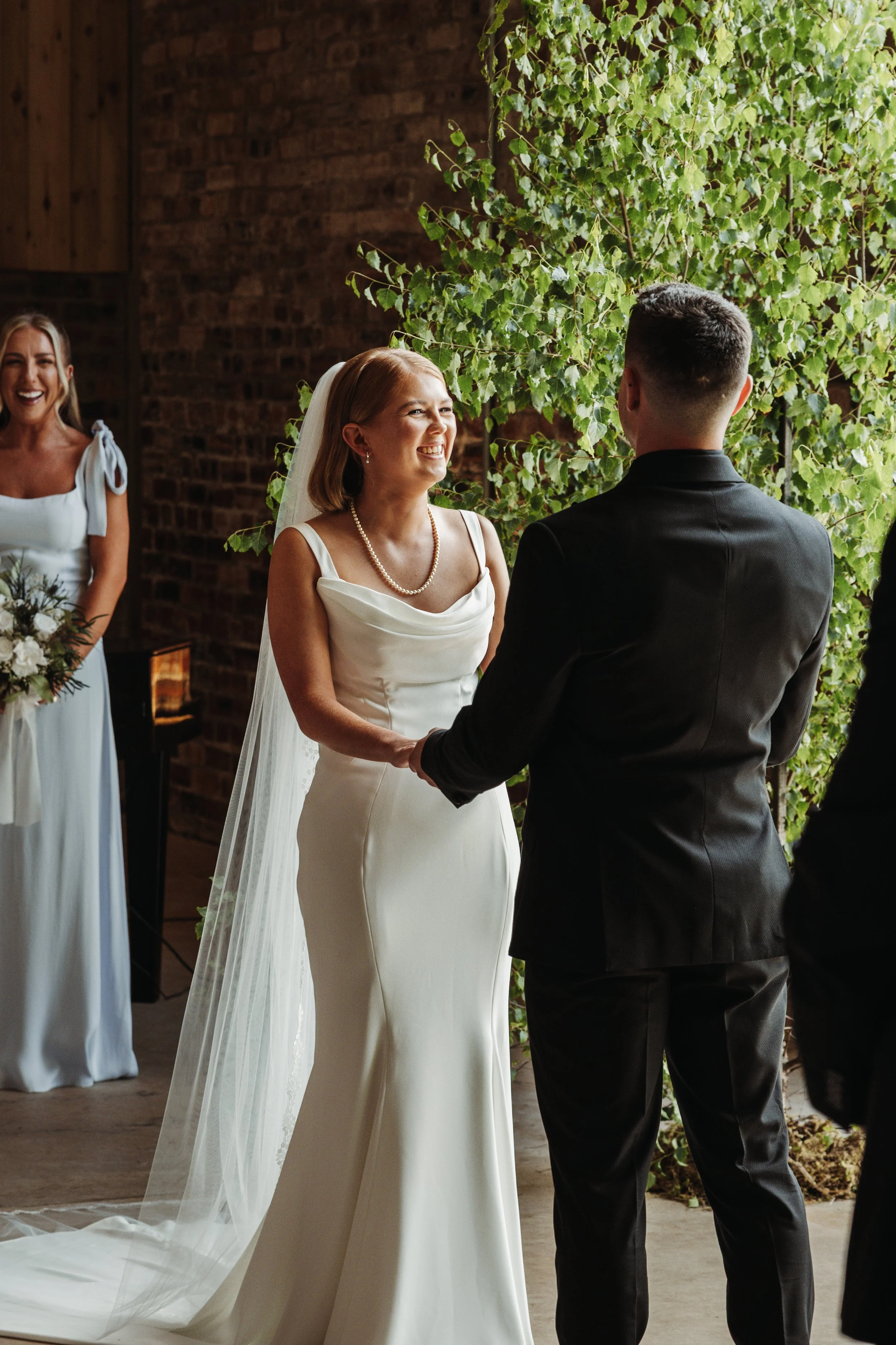 A wedding ceremony with a bride and groom exchanging vows. The bride is smiling, wearing a white wedding dress, pearl necklace, and veil. The groom, dressed in a black suit, is facing her. Bridesmaid, in a white dress, is standing with a bouquet, wat