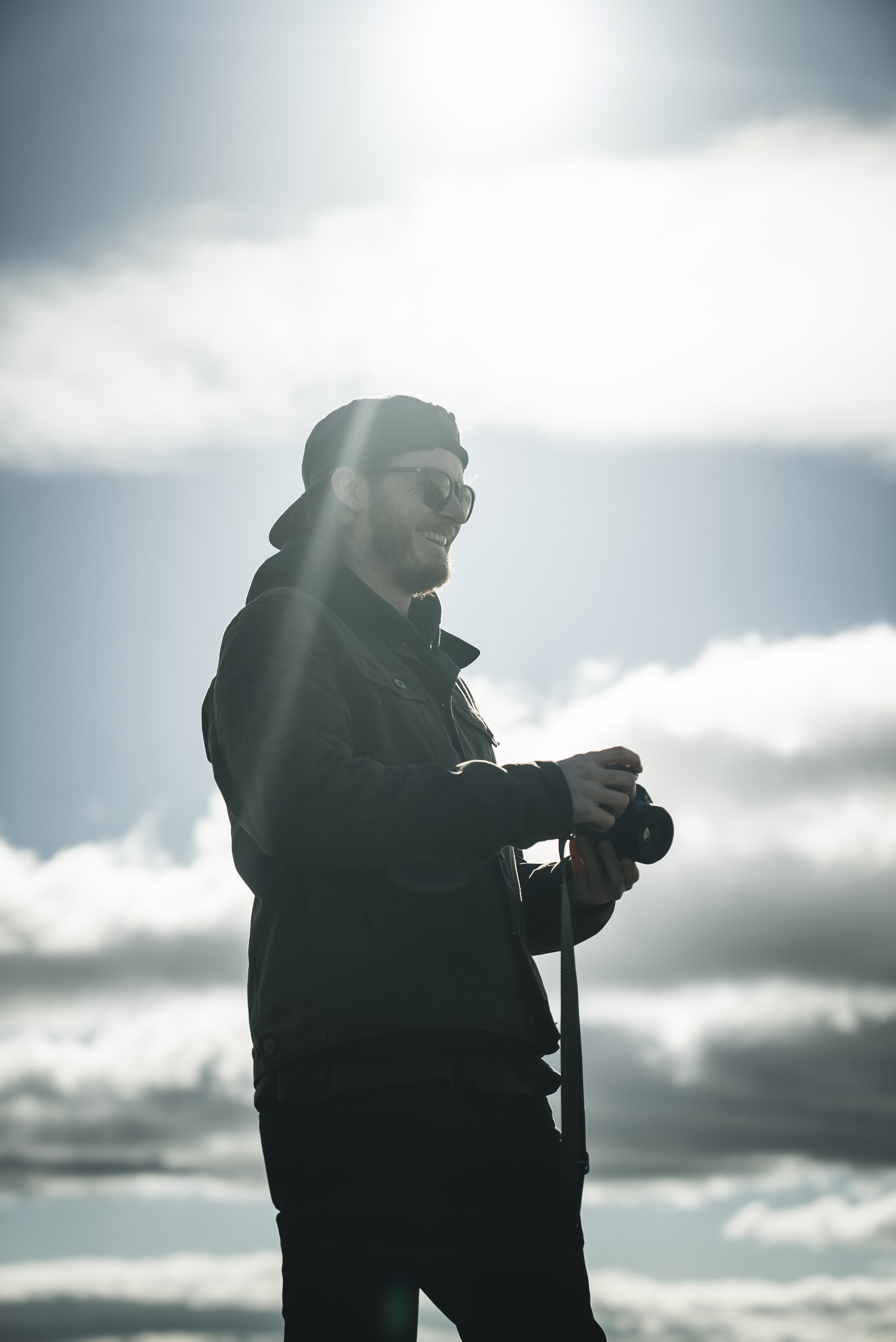Ruaridh Kidd from RKidd Weddings wearing sunglasses and a black jacket, smiling and holding a camera, standing outdoors against a background of cloudy skies and sunlight.