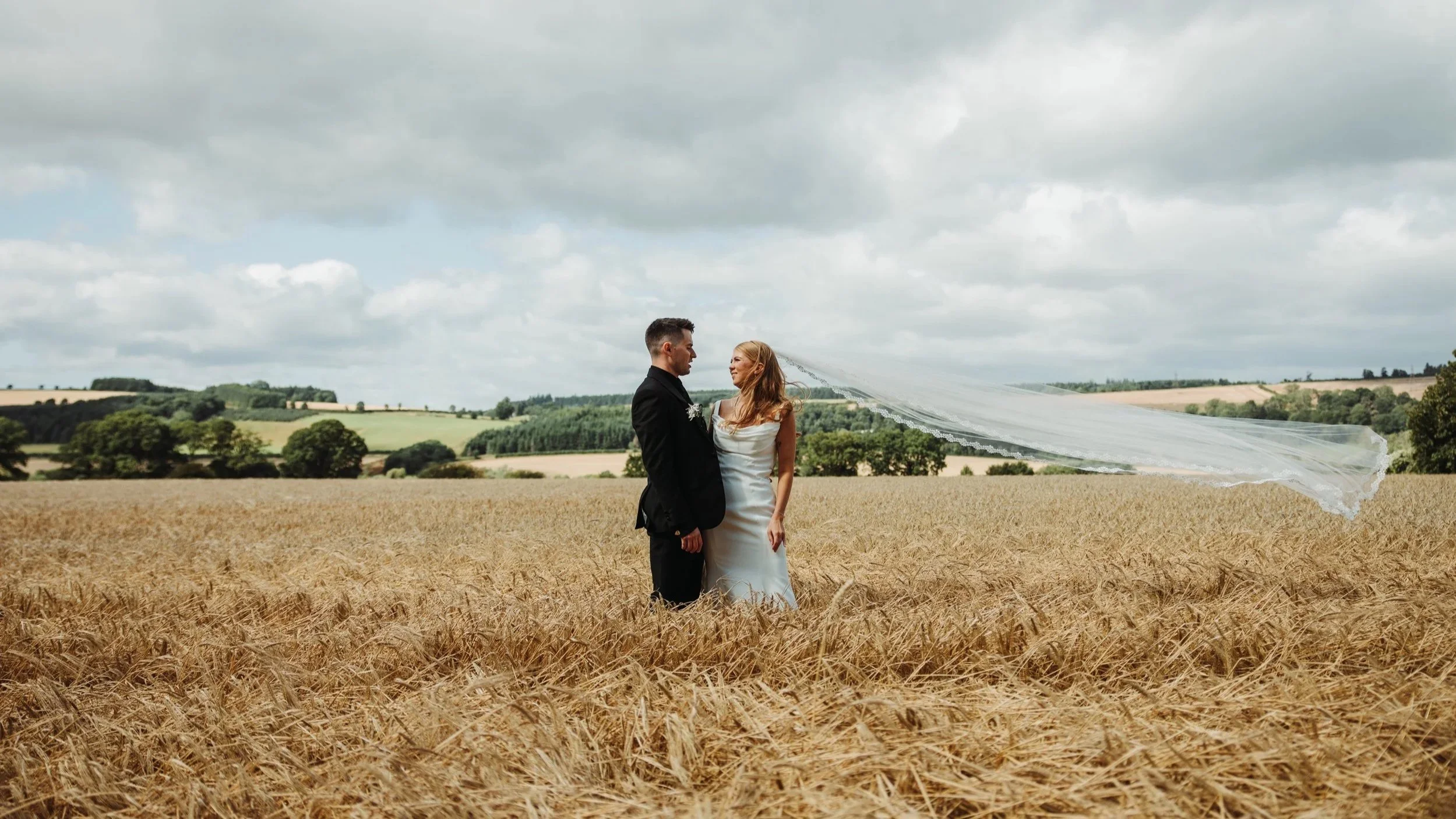 A bride and groom standing in a wheat field under a cloudy sky, with a woman in a wedding dress and veil standing next to the groom.