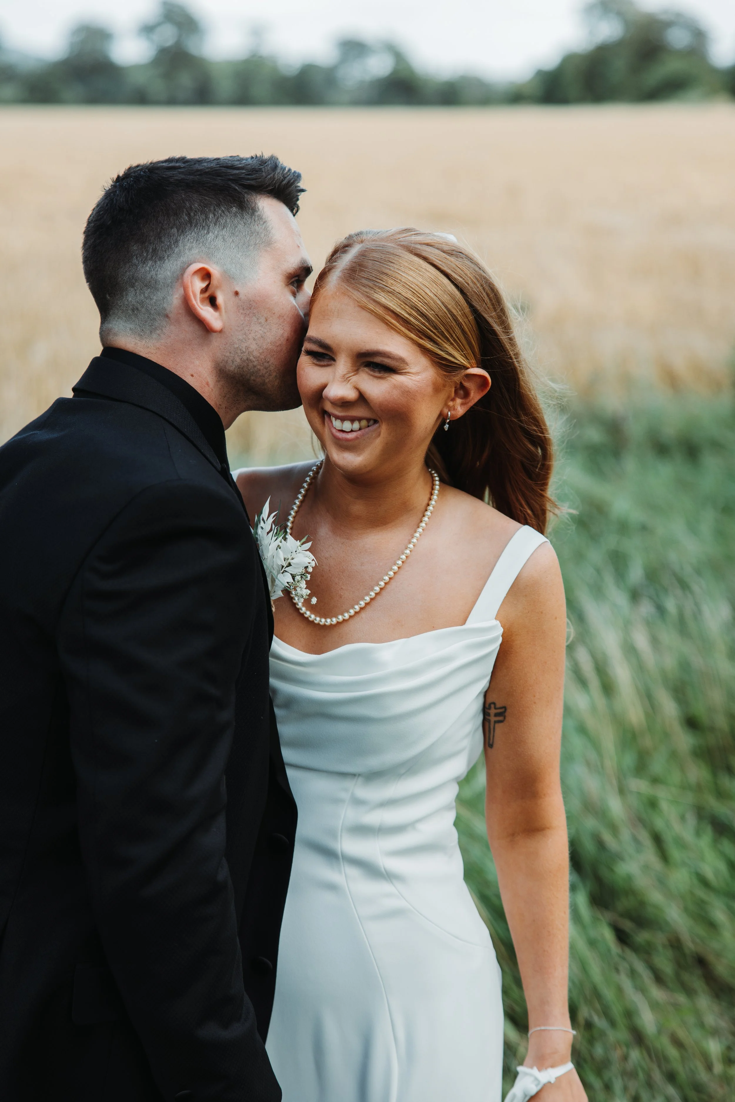 A man in a black suit kisses a smiling woman in a white dress, standing outdoors with a blurred field and trees in the background.