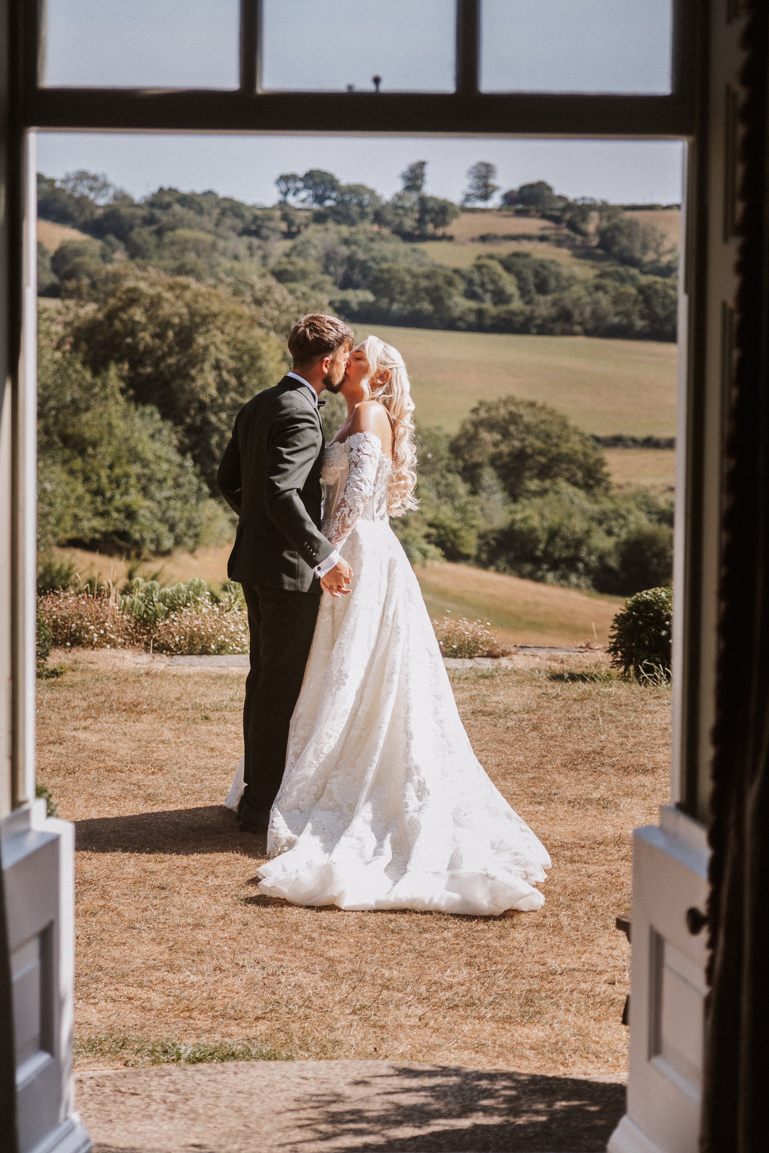 A bride and groom sharing a kiss outside, framed by an open door with white shutters, on a grassy hill with trees and rolling hills in the background.