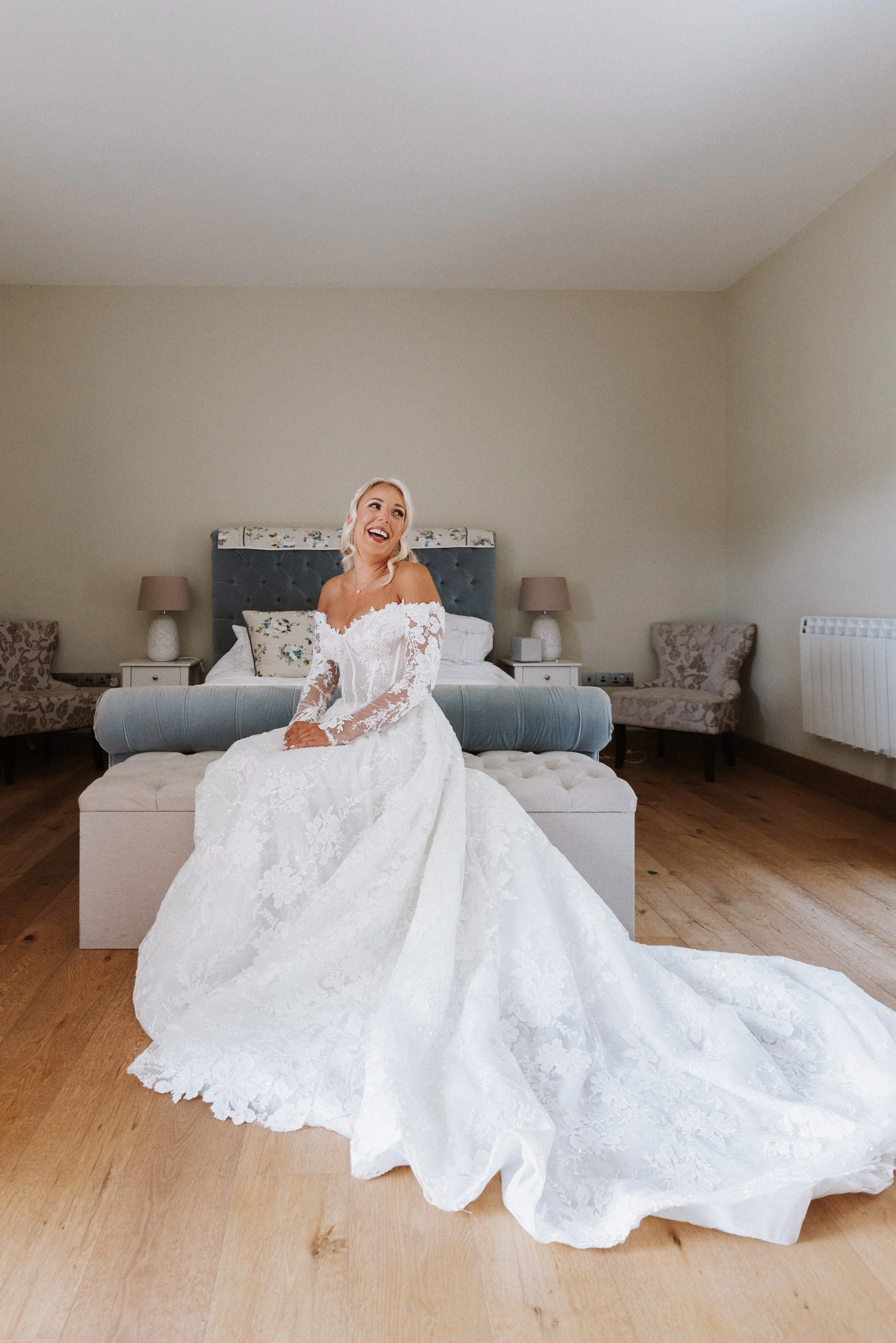 A woman in a white lace wedding gown sitting on a cushioned ottoman in a bedroom, smiling and looking to her right. The bedroom has beige walls, two nightstands with lamps, and two chairs with patterned upholstery.