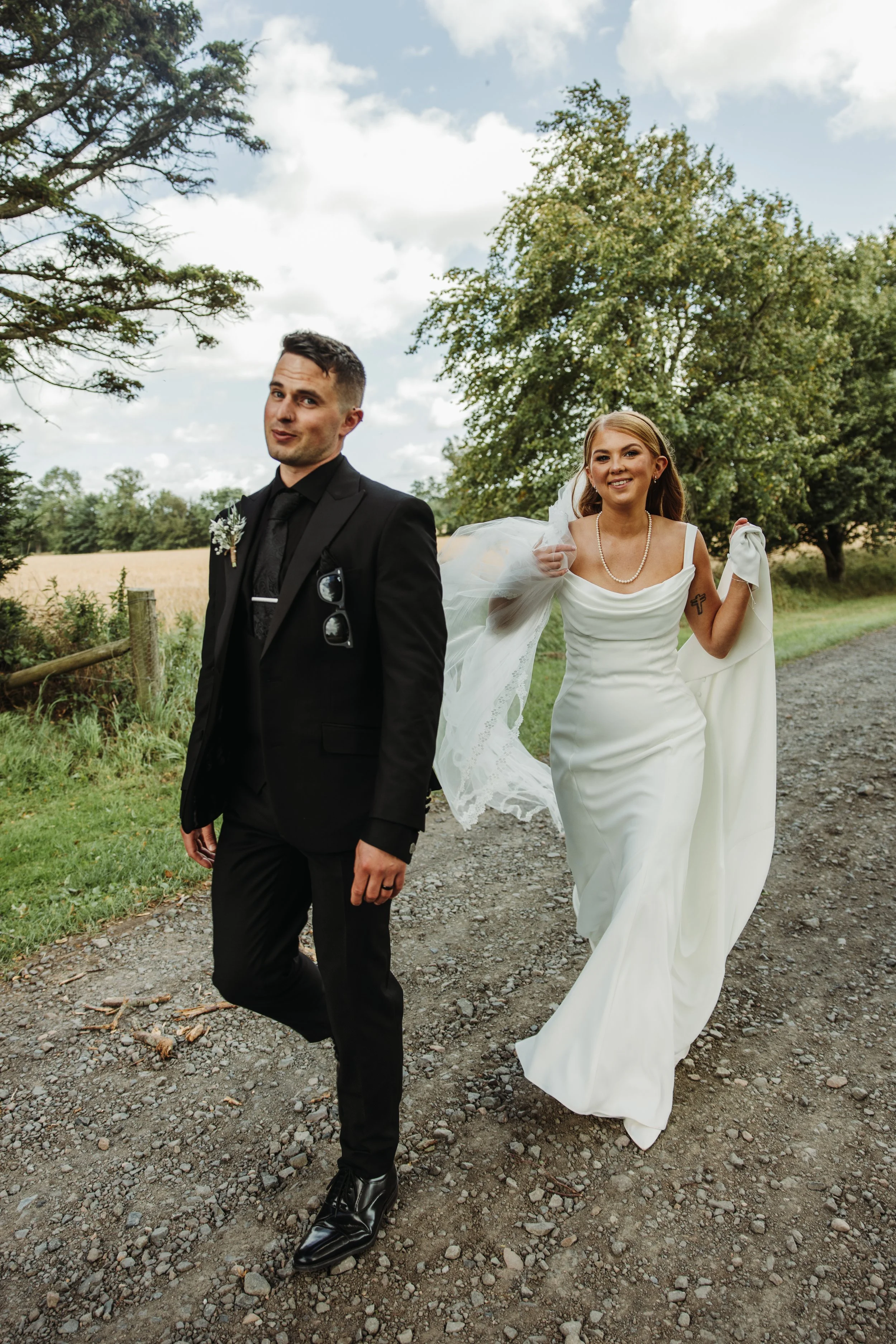 A bride and groom walking outdoors on a gravel path, with trees and fields in the background. The groom is wearing a black suit and tie, and the bride is in a white wedding gown, holding her dress and smiling.