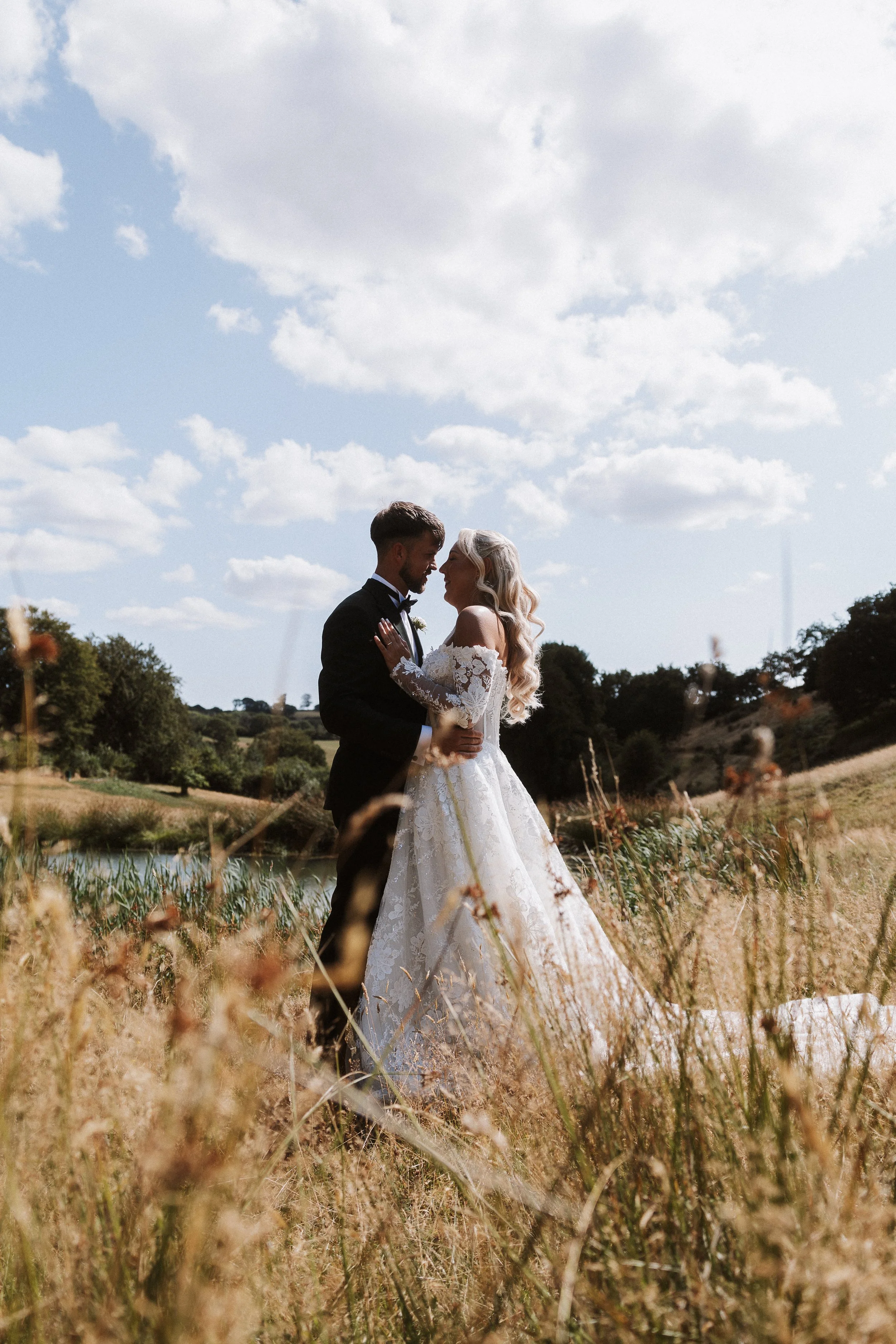 A newlywed couple embraces outdoors during their wedding photoshoot, with tall grass in the foreground and a partly cloudy sky overhead.