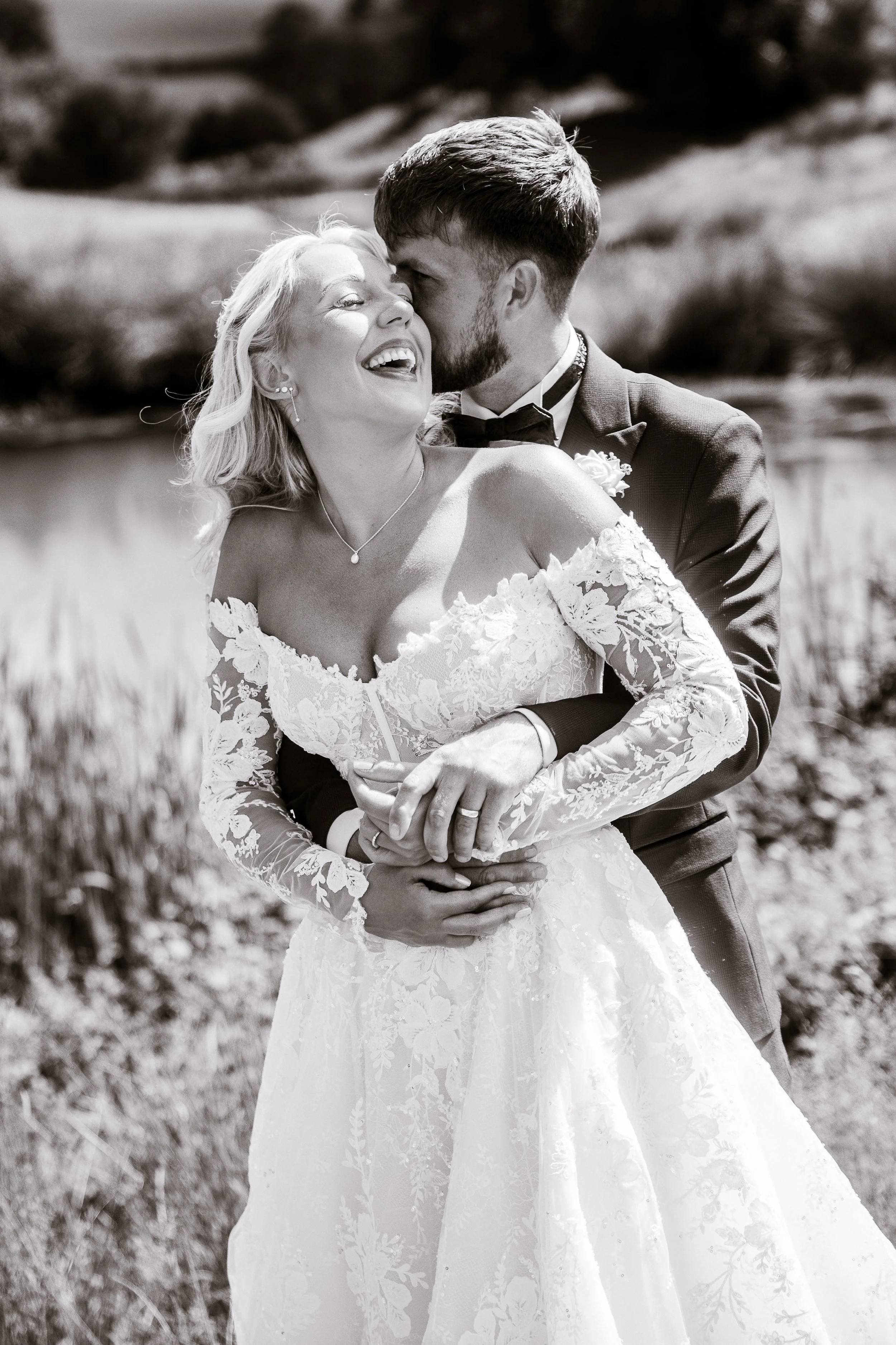 A black-and-white photo of a woman and a man at a wedding. The woman, in a lace wedding dress, is laughing with her eyes closed, while the man, in a tuxedo, kisses her on the cheek from behind. They are outdoors in a field.