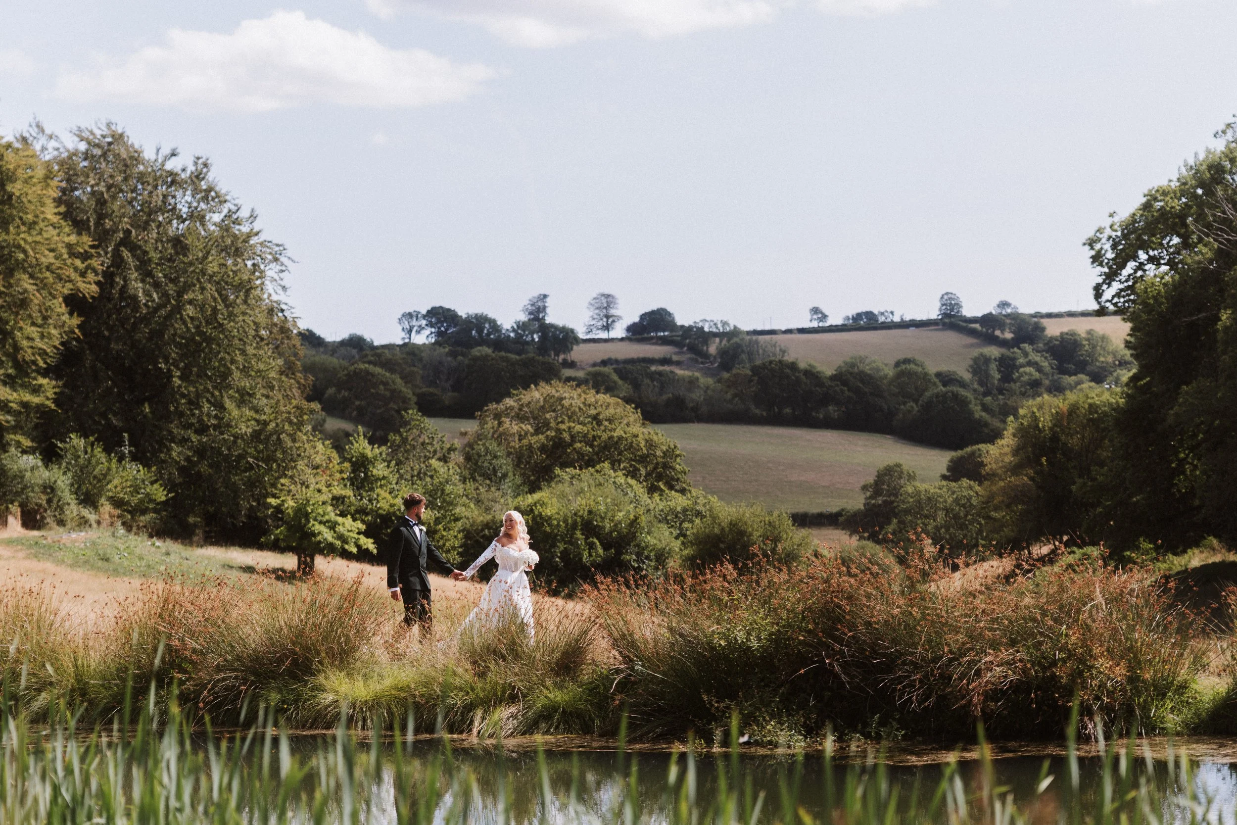 A bride and groom holding hands while walking through a scenic field with trees and hills in the background.