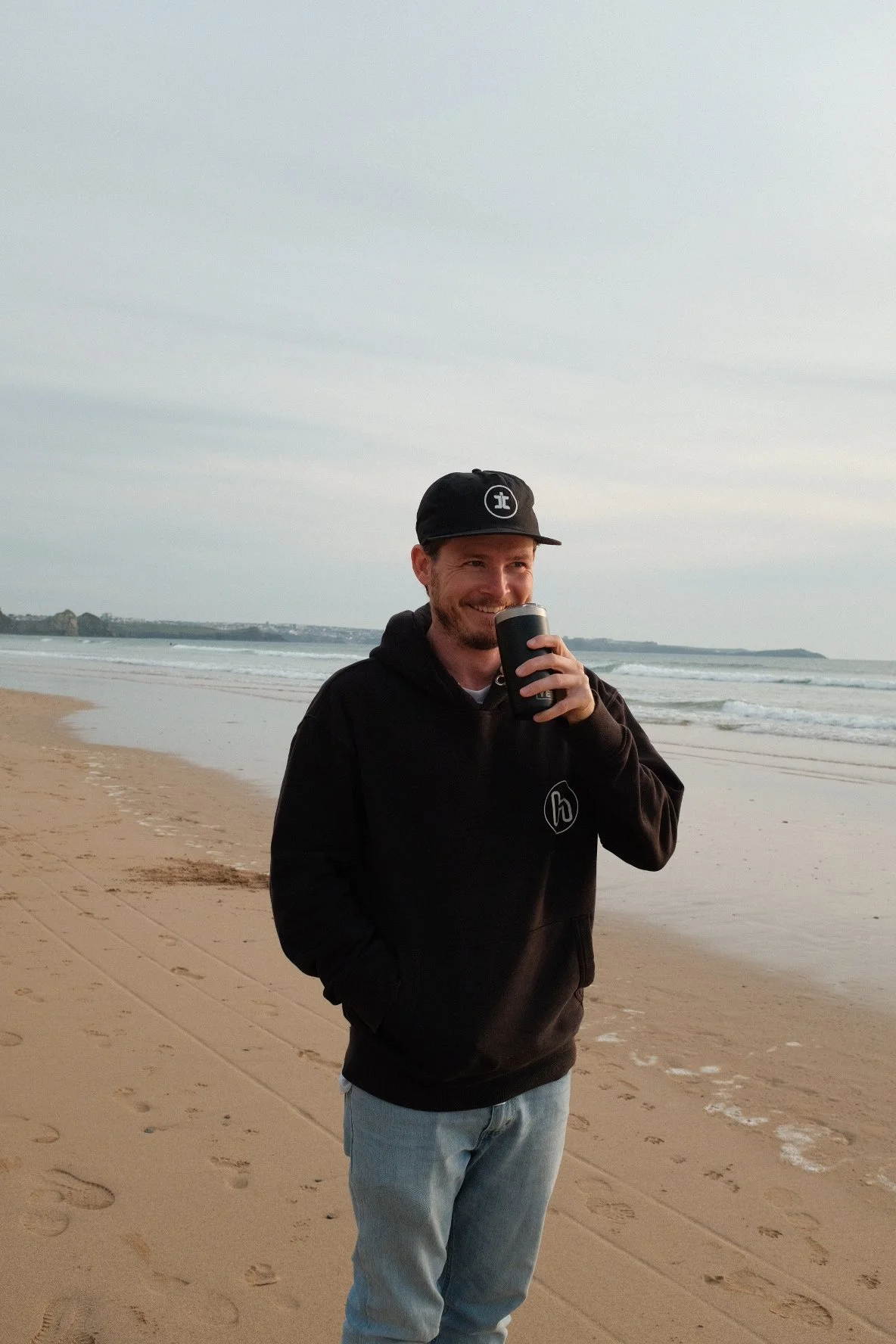 Ruaridh Kidd from RKidd Weddings is standing on a sandy beach, smiling and holding a black tumbler, with the ocean and cloudy sky in the background.