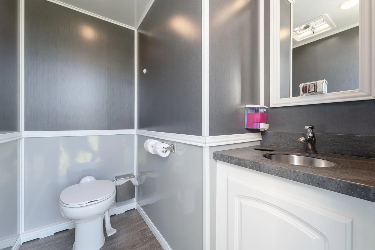 A small bathroom with gray and white walls, a white toilet, a gray countertop with an oval sink, a mirror, a purple soap dispenser, and a small window on the ceiling.