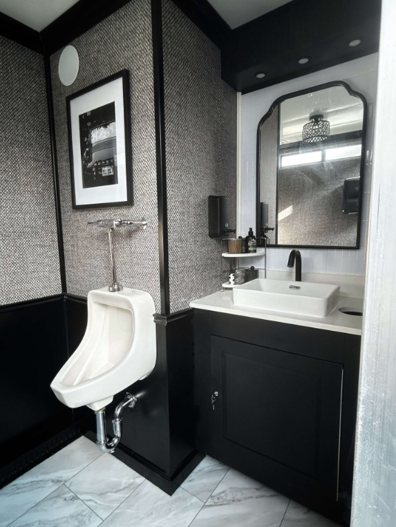 A modern bathroom with a white sink, black cabinet, and a large mirror. There is a urinal next to the sink and a framed black-and-white photograph on the wall.