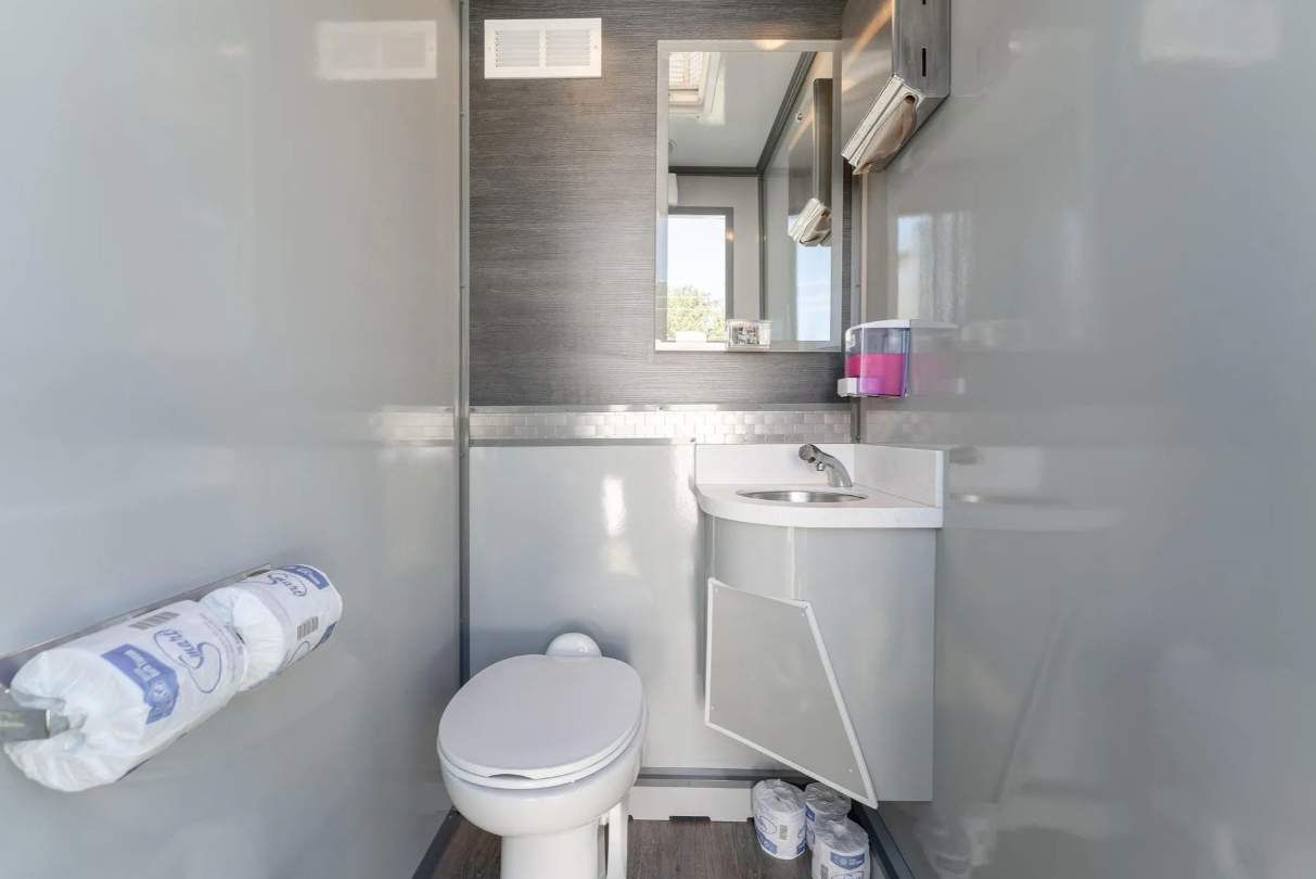 Small, compact bathroom with a toilet, a small sink, and a mirror, in a portable or mobile unit. Toilet paper rolls are on a holder, and soap dispenser and paper towel dispenser are on the wall.