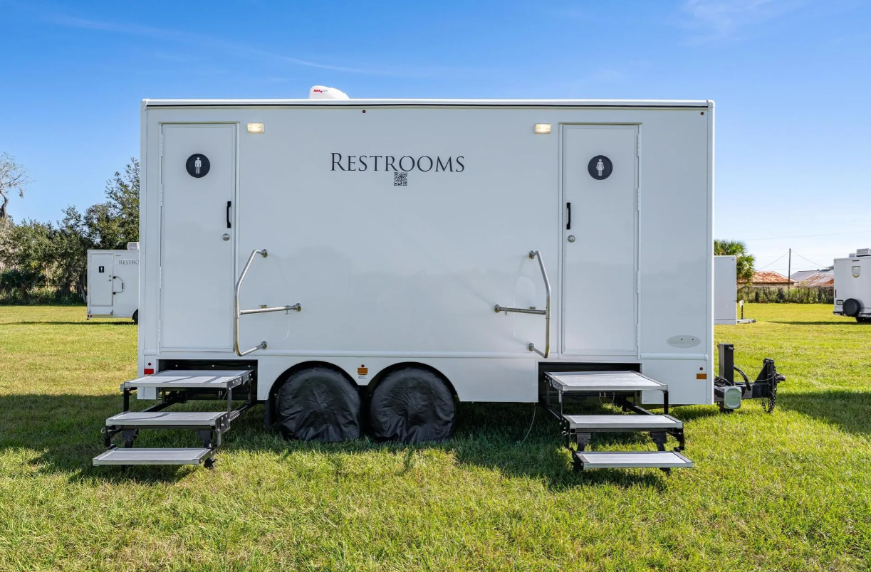White portable restroom trailer with two doors, each marked with a male or female symbol, situated on a grassy field with smaller trailers in the background under a clear blue sky.
