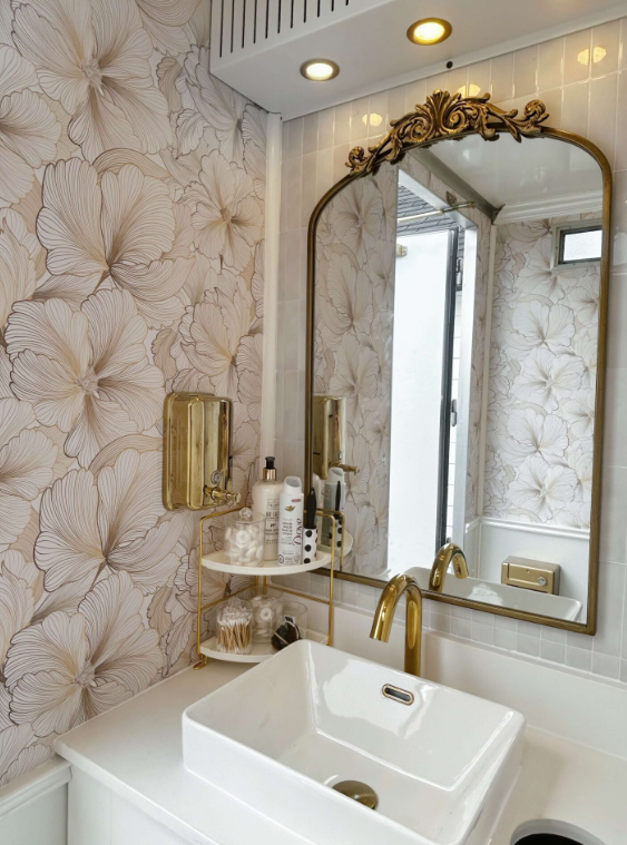 Bathroom vanity with a white square sink, gold faucet, large mirror with ornate gold frame, wallpaper with large floral patterns, and a small round shelf holding toiletries, with window and exterior door reflected in the mirror.