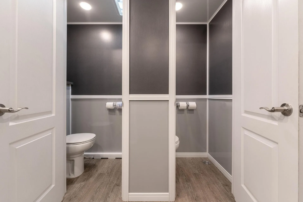 Two public restroom stalls with white doors, interior walls painted in dark and light gray, and a toilet inside each stall.