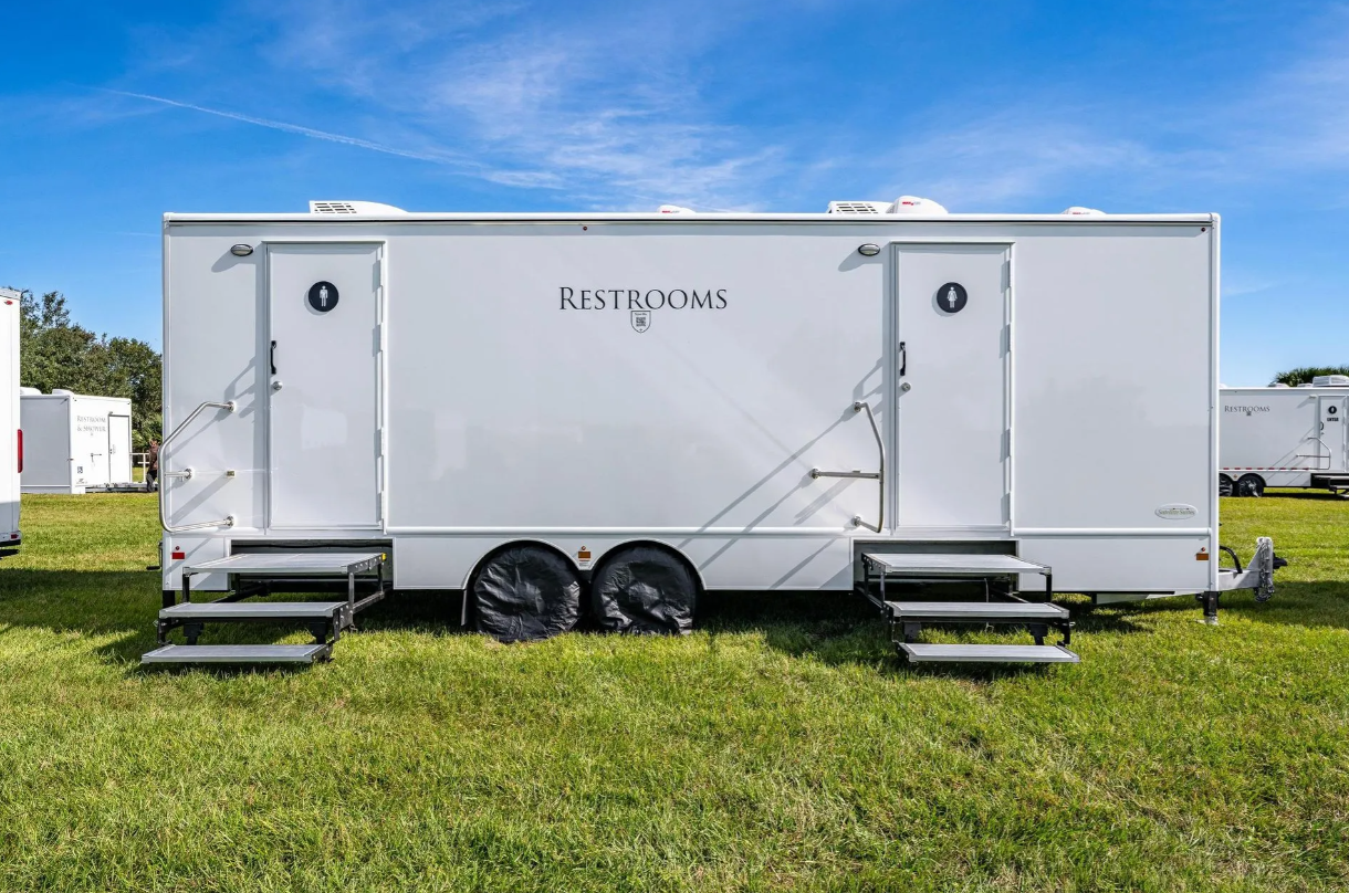 A portable restroom trailer with two entry doors, stairs, and signage indicating men's and women's restrooms, situated on a grassy field with other trailers in the background.