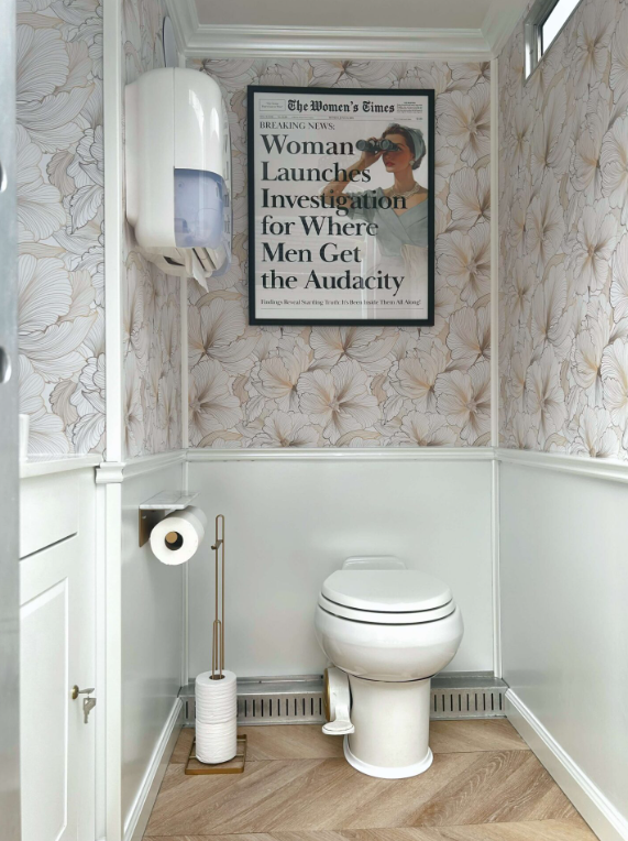 A small bathroom with a white toilet, a toilet paper holder with roll, a framed newspaper headline on the wall, patterned wallpaper, a wall-mounted hand dryer, and a small window near the ceiling.