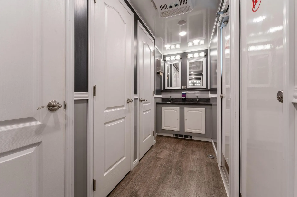 A small bathroom with white doors and cabinets, a black countertop with two sinks, and a large mirror above each sink. There are multiple light fixtures above the mirrors, a ceiling vent, and wood-look flooring.
