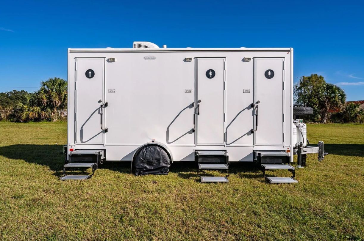 White portable restroom trailer with four doors, each with a male or female sign, set on grassy field with blue sky and trees in the background.