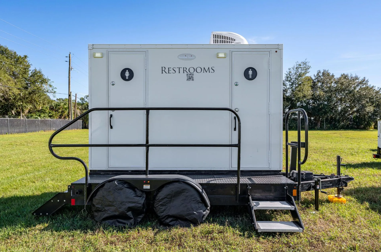 A mobile restroom trailer with two doors, one for men and one for women, situated on a grassy field under a clear sky.