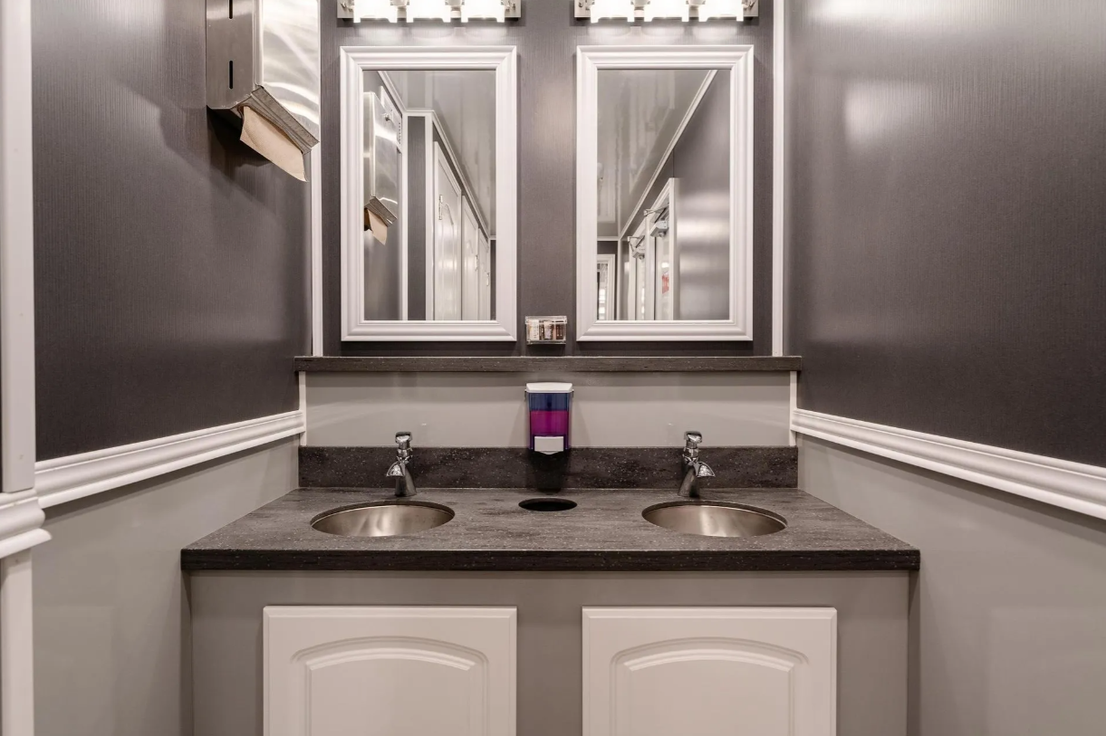 A bathroom sink area with two stainless steel sinks, a large mirror, and a soap dispenser in the middle. The walls are painted gray with white trim, and there are two small mirrors above the sinks.