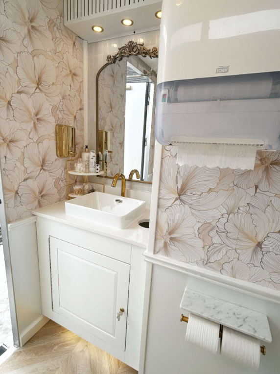 A bathroom with floral wallpaper, a white vanity with a square sink, a gold faucet, a large decorative mirror, a soap dispenser, and a wall-mounted paper towel dispenser. There is a window in the reflection of the mirror and ceiling recessed lighting