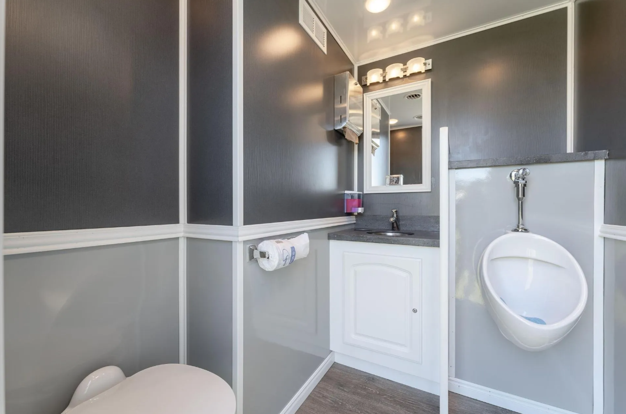 A small bathroom with a dark gray wall and white trim, a urinal, a sink with a small countertop, a mirror, a wall-mounted paper towel dispenser, a mirror above the sink, and a wall-mounted hand dryer.