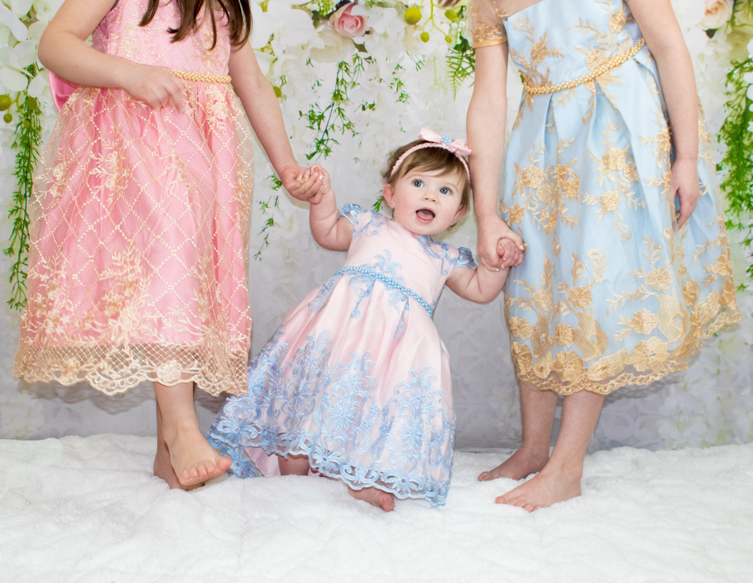 A young girl wearing a pink dress with blue embroidery is holding hands with two women wearing light blue and pink dresses with gold embroidery, standing on a white fluffy surface in front of a floral backdrop.