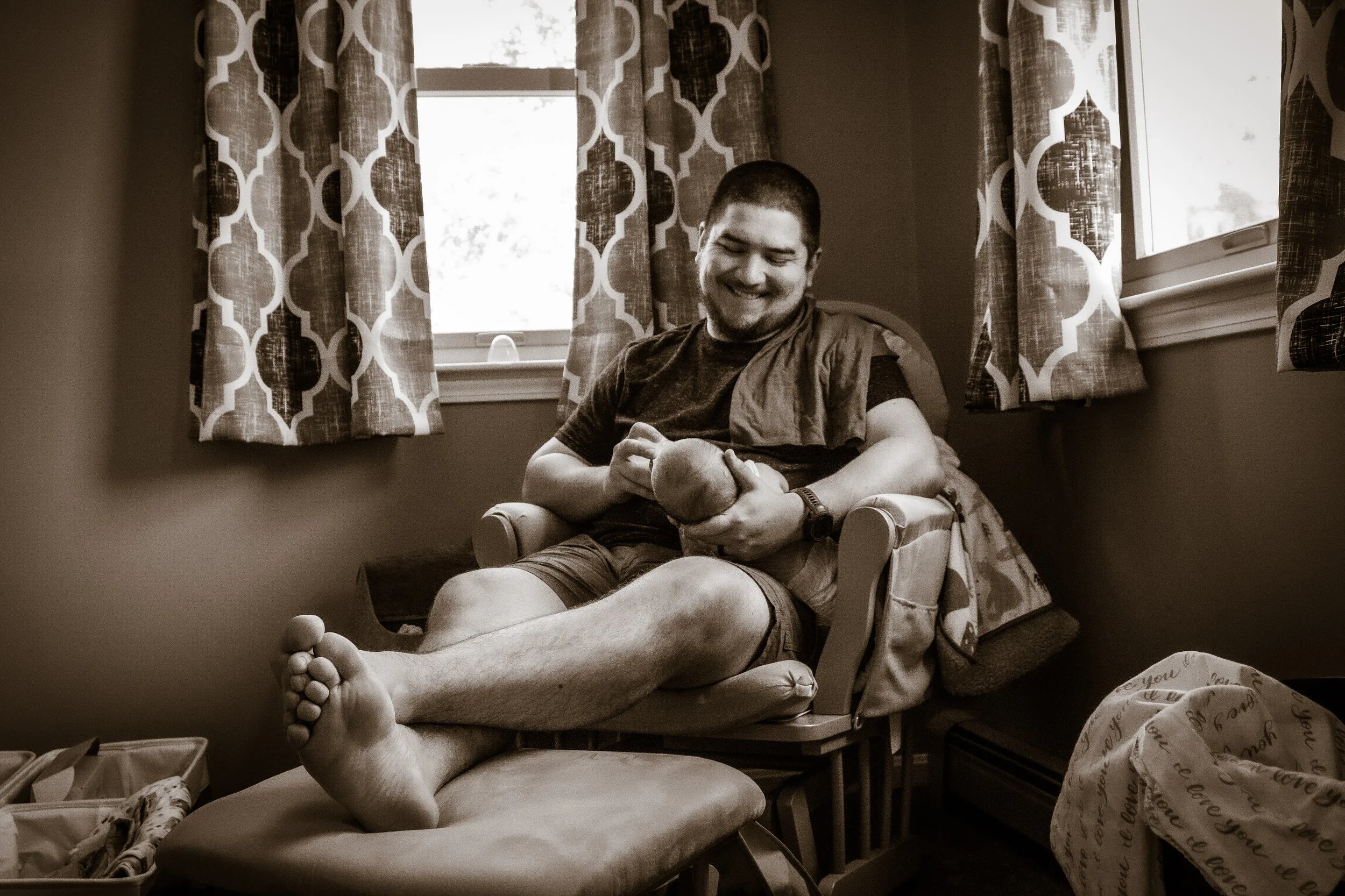 A man sitting in a chair, smiling, holding a newborn baby in his arms, in a room with a window and patterned curtains.