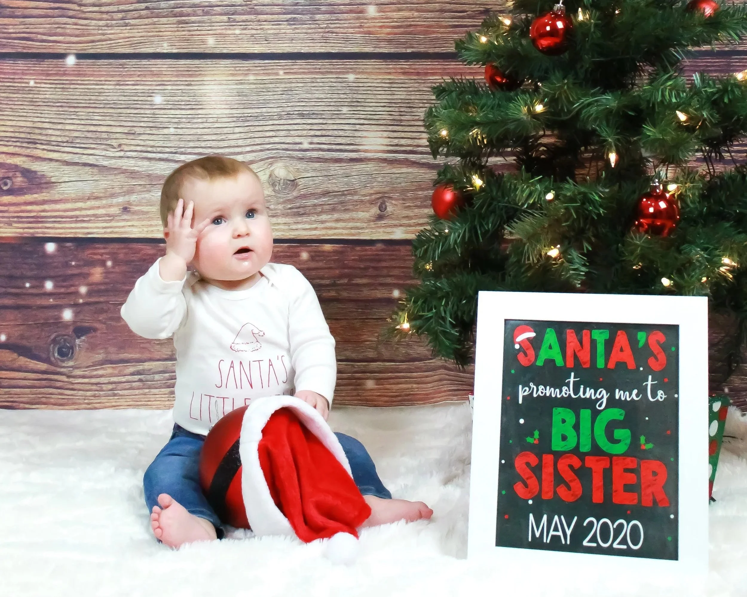 A young child sitting on a plush white surface near a Christmas tree decorated with red ornaments and lights. The child is wearing a white shirt with the message "Santa's little" on it. There is a sign on the right with colorful holiday writing, mentioning an event in May 2020.