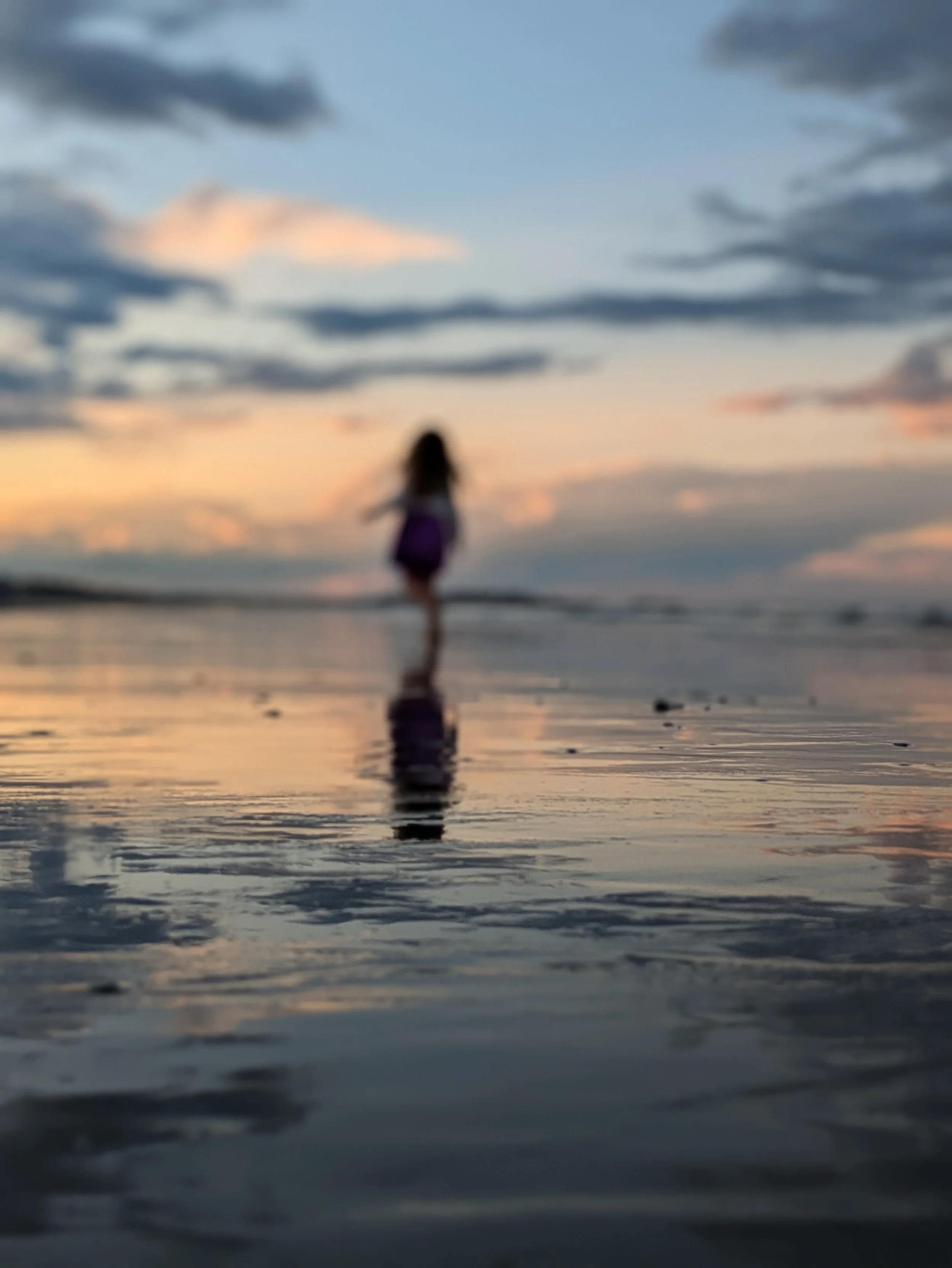A blurry image of a person walking on the beach during sunset, with clouds in the sky and the reflection of the sky on the wet sand.