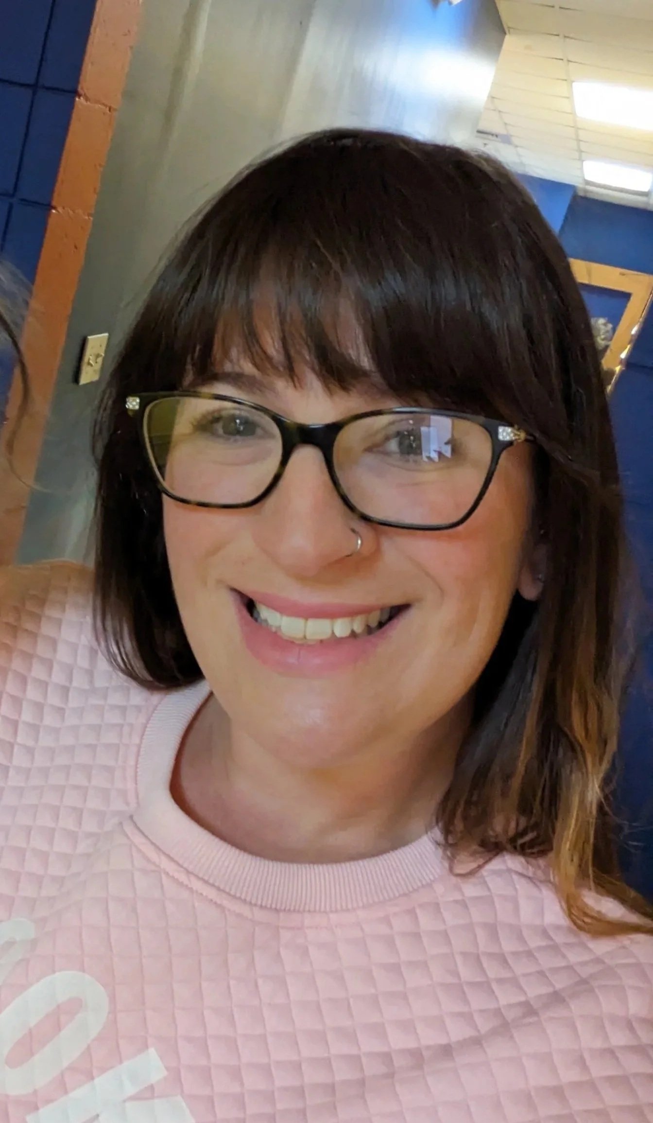 A woman with dark brown hair, glasses, and a nose piercing smiling for a selfie inside a building with wooden and blue walls.
