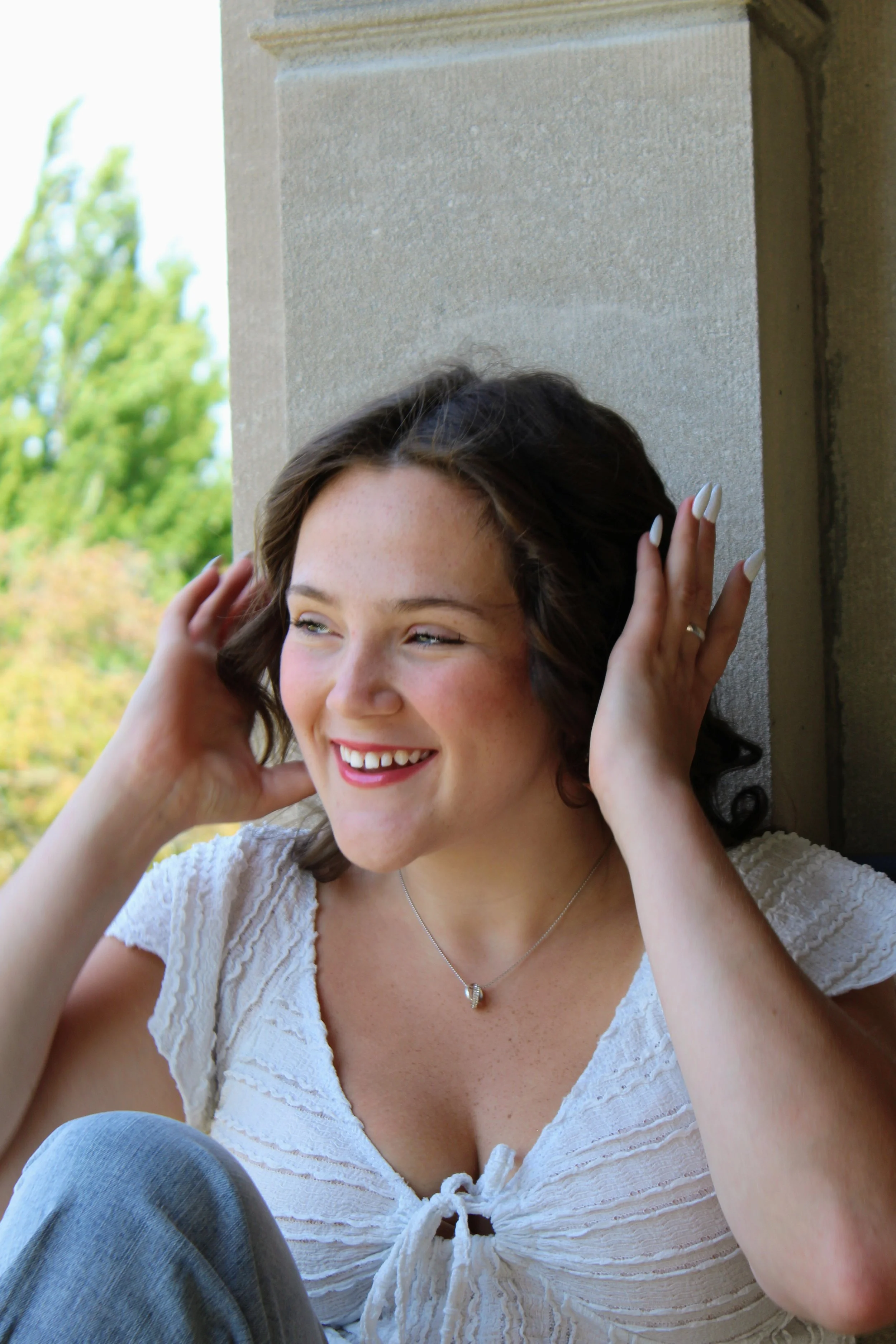 A woman with curly brown hair smiling, wearing a white lace blouse with a front tie, and a delicate necklace, sitting outdoors against a stone wall with green trees in the background.