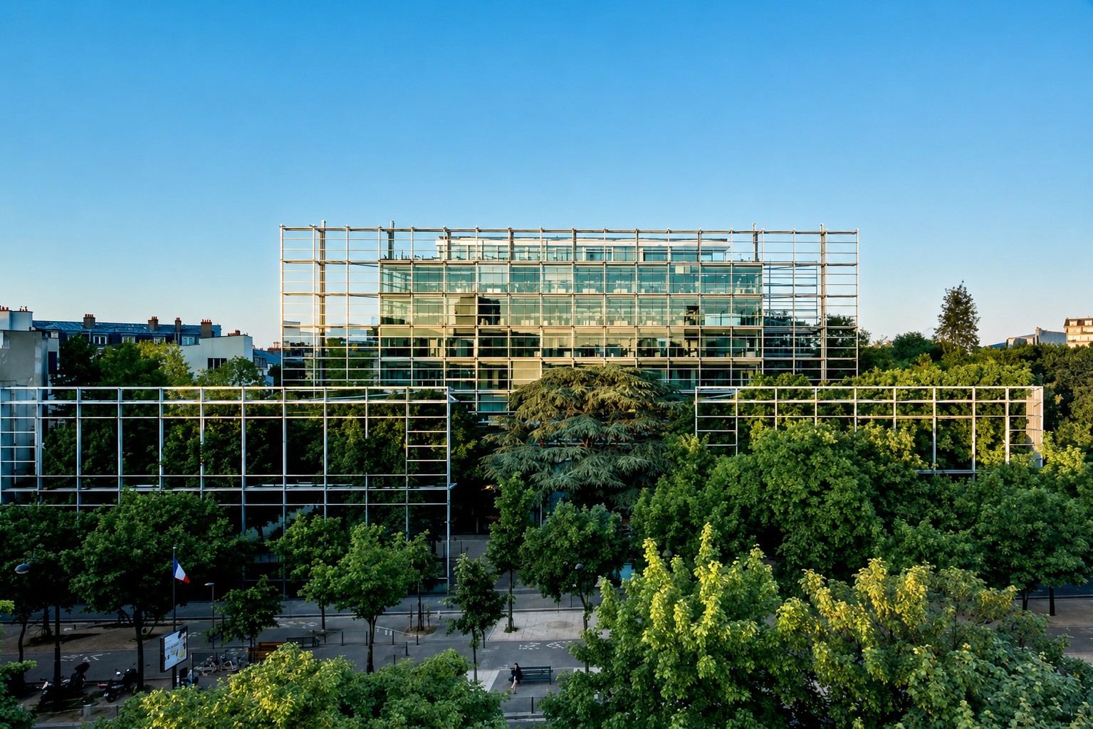 Bâtiment moderne en verre entouré d'arbres verts et d'un ciel clair, avec une structure métallique en premier plan.