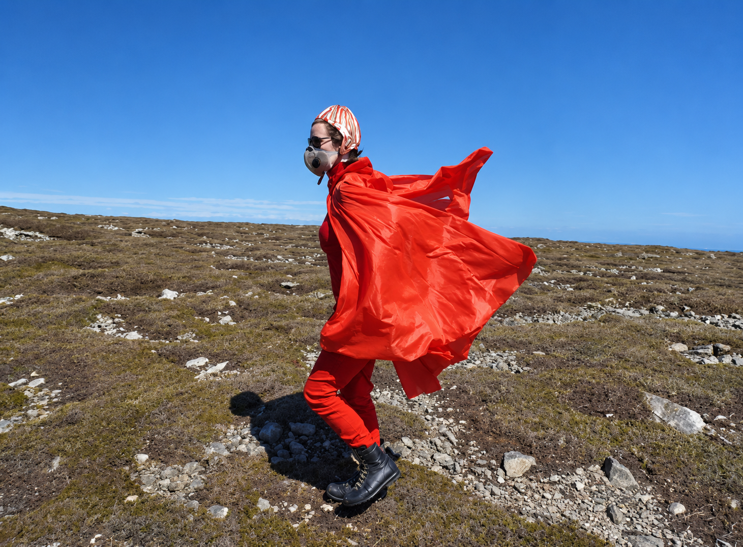 Personne en plein air portant un vêtement rouge, un masque et des bottes, avec un ciel bleu clair.