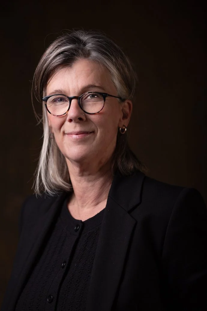 Portrait of a middle-aged woman with shoulder-length gray hair, wearing glasses, a black blazer, and a black top, against a dark background.