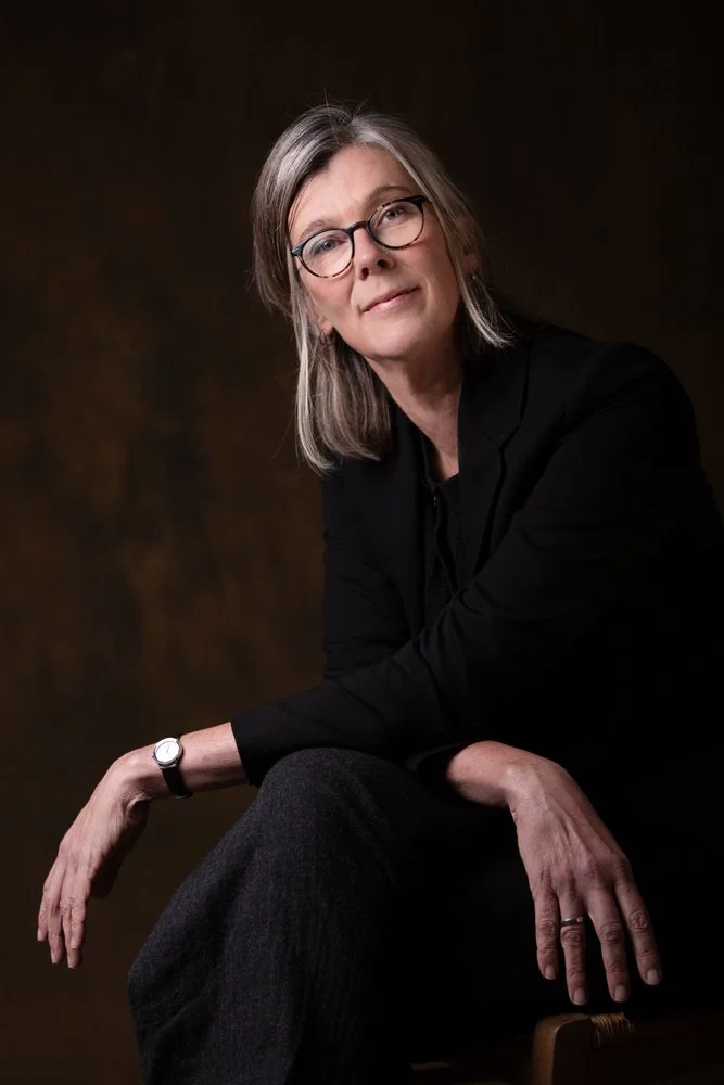 A middle-aged woman with gray hair and glasses sitting in a black blazer and dark pants against a dark brown background.