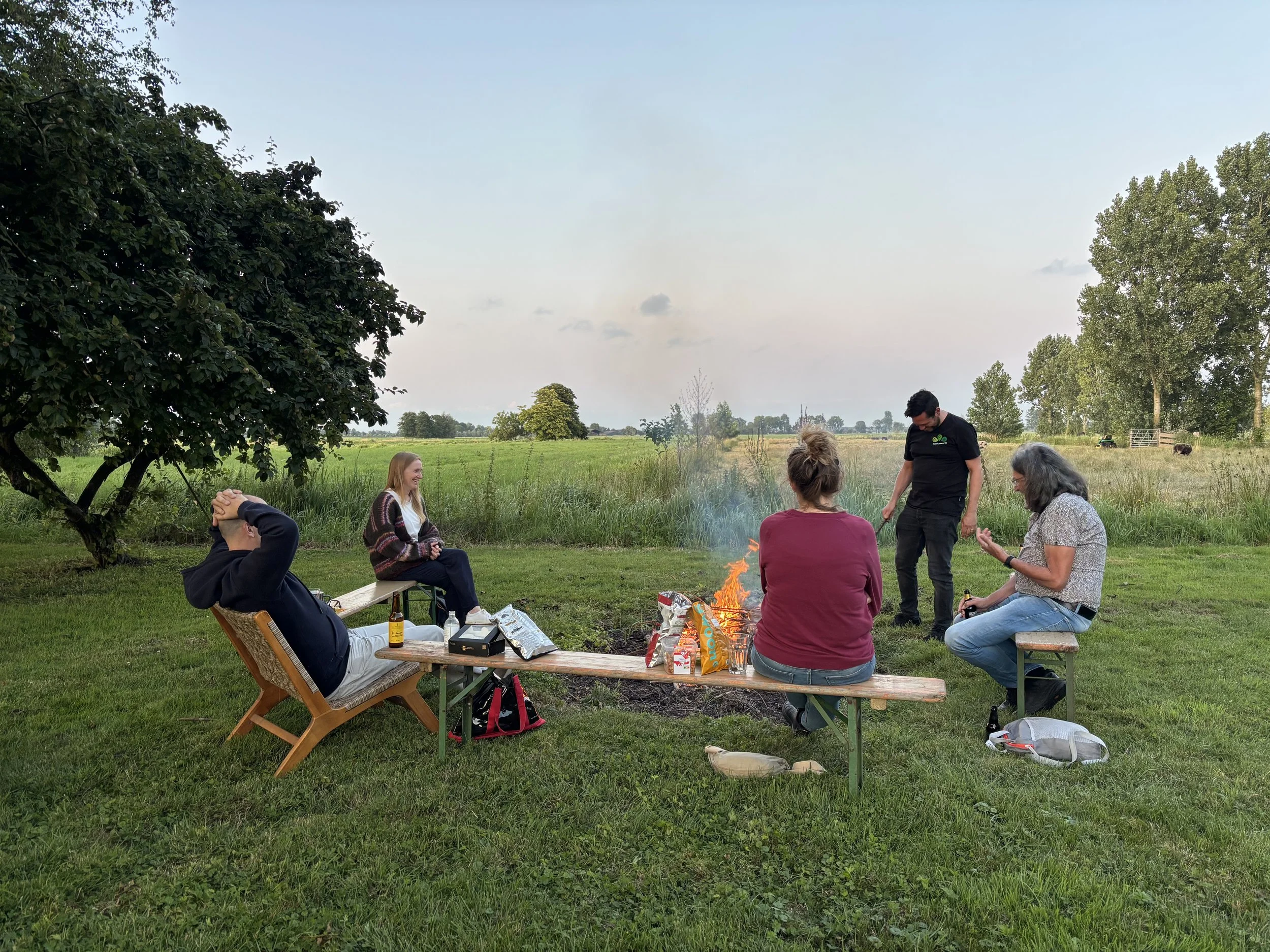Group of five people gathering outdoors around a campfire in a grassy field, with trees and an open sky in the background, some sitting on benches and others standing, enjoying a social moment with snacks and drinks.