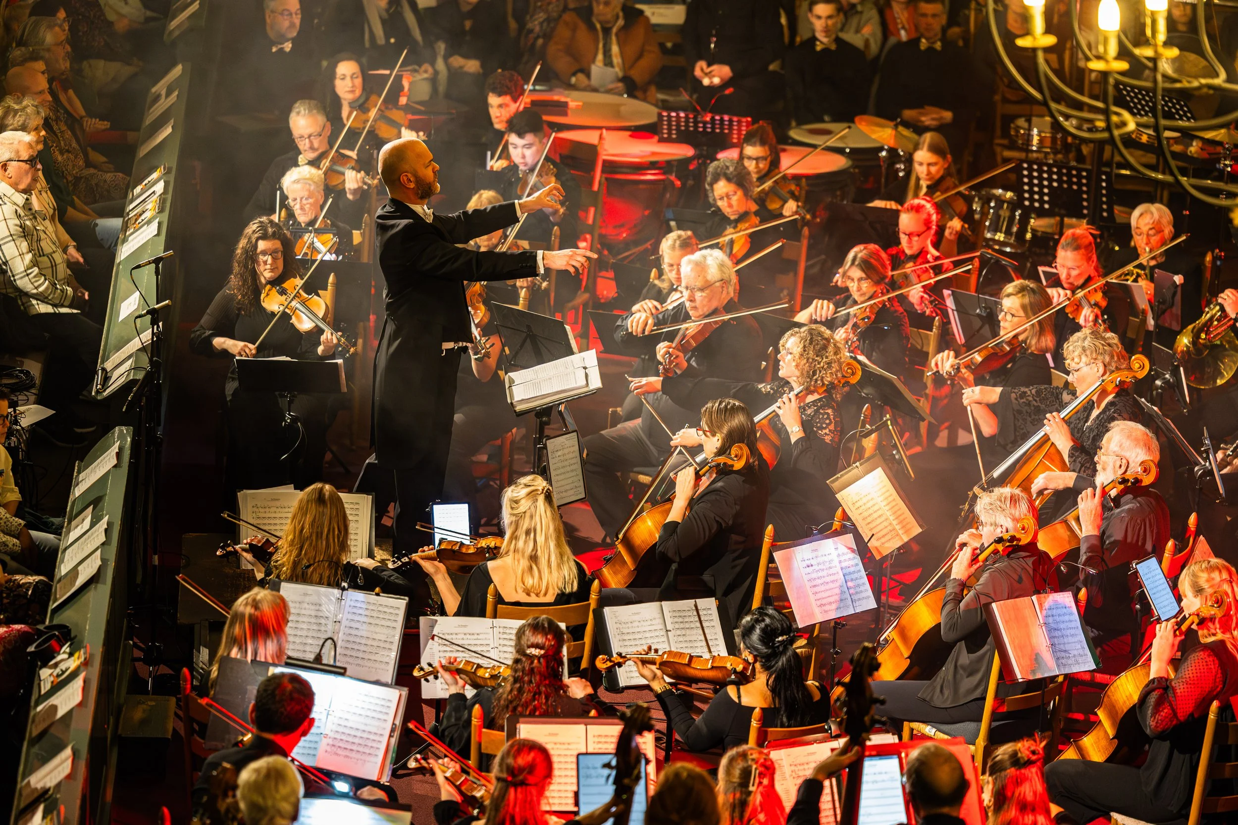 A symphony orchestra performing on stage with the conductor in the center, surrounded by musicians playing violins, cellos, and other instruments, illuminated by warm stage lighting.