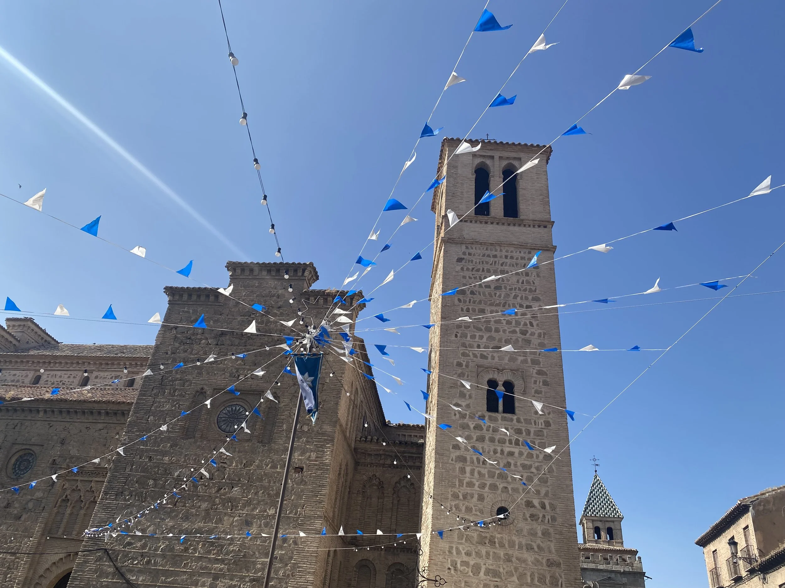 A historic stone church with tall towers, decorated with blue and white triangular flags hanging in the air against a clear blue sky.