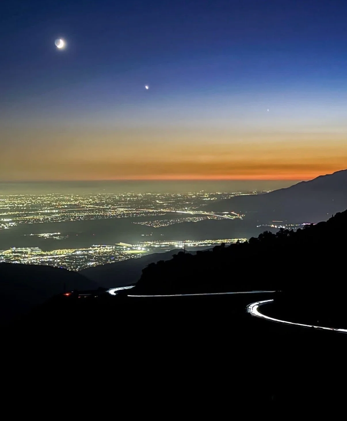 Nighttime view of city lights from a mountain, with a winding road in the foreground, and a sunset with a crescent moon and planets in the sky.