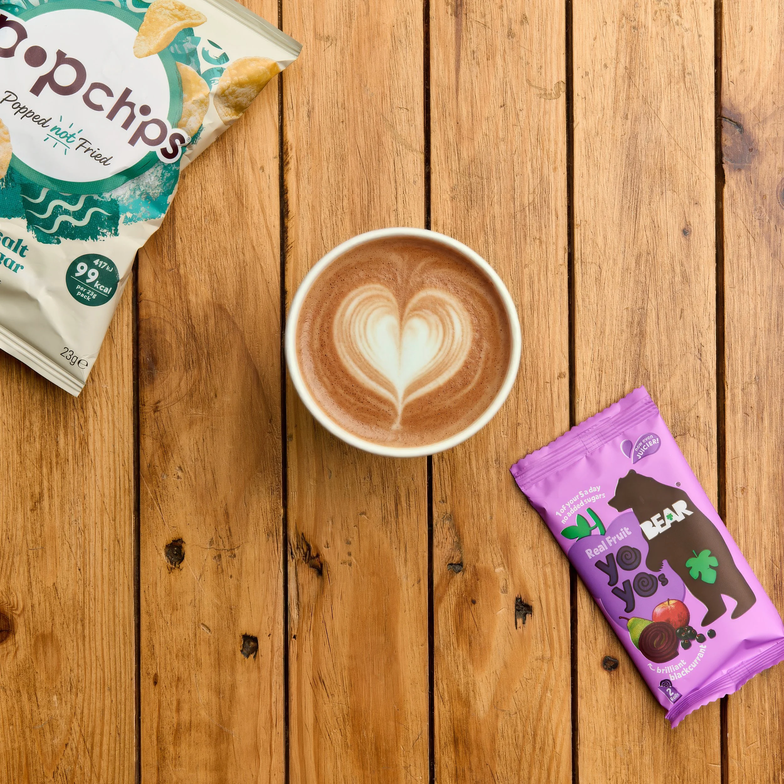 A partially visible bag of popcorn chips, a cup of coffee with foam art, and a purple packet of bear yoyo on a wooden surface.
