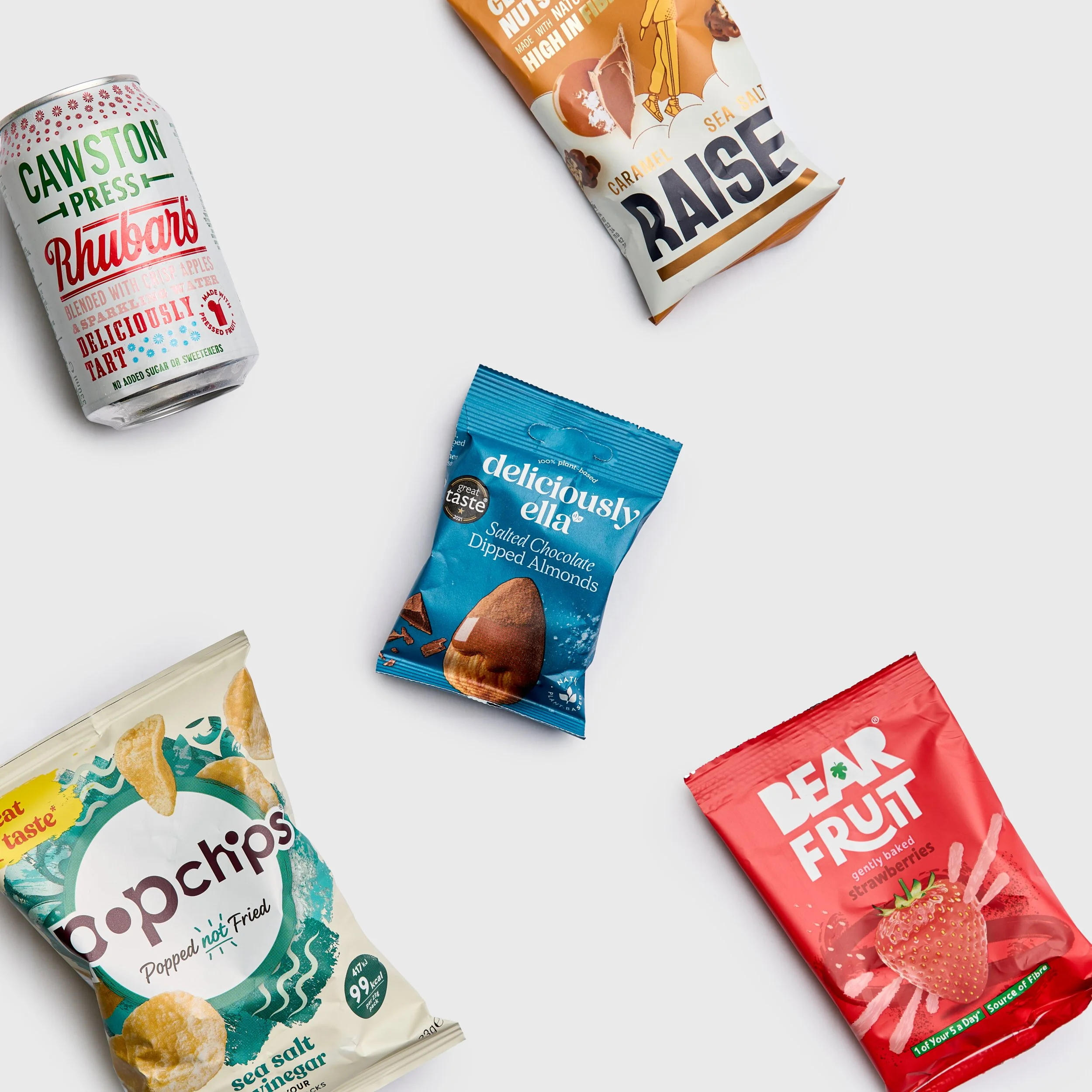 Arrangement of snack packages on a white surface, including a can of Cawston Press sparkling apple water, a blue packet of deliciou sally salted chocolate dipped almonds, an orange and white raise, and a red package bear fruit