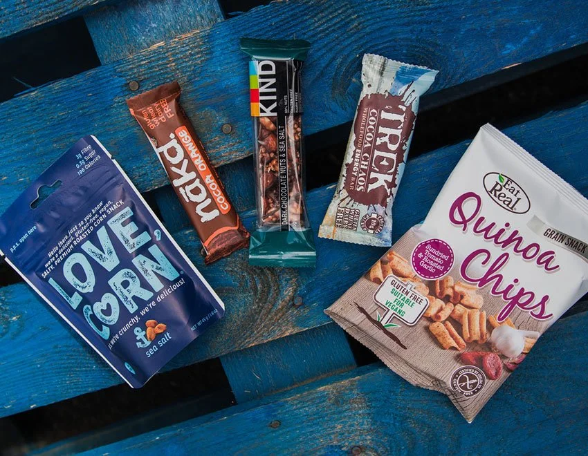 Assorted snack bars including kind and naked and a bag of quinoa chips on a blue wooden surface.