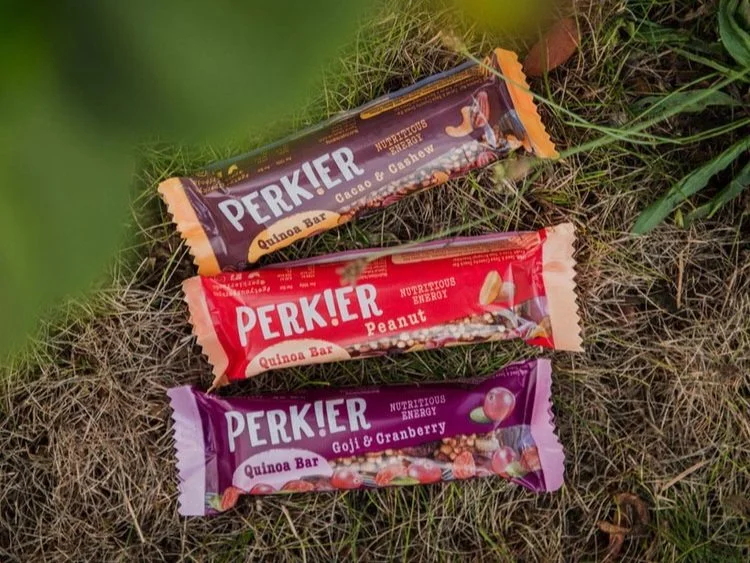 Three Perkier quinoa bar packages in different flavors lying on grass and dirt outdoors.