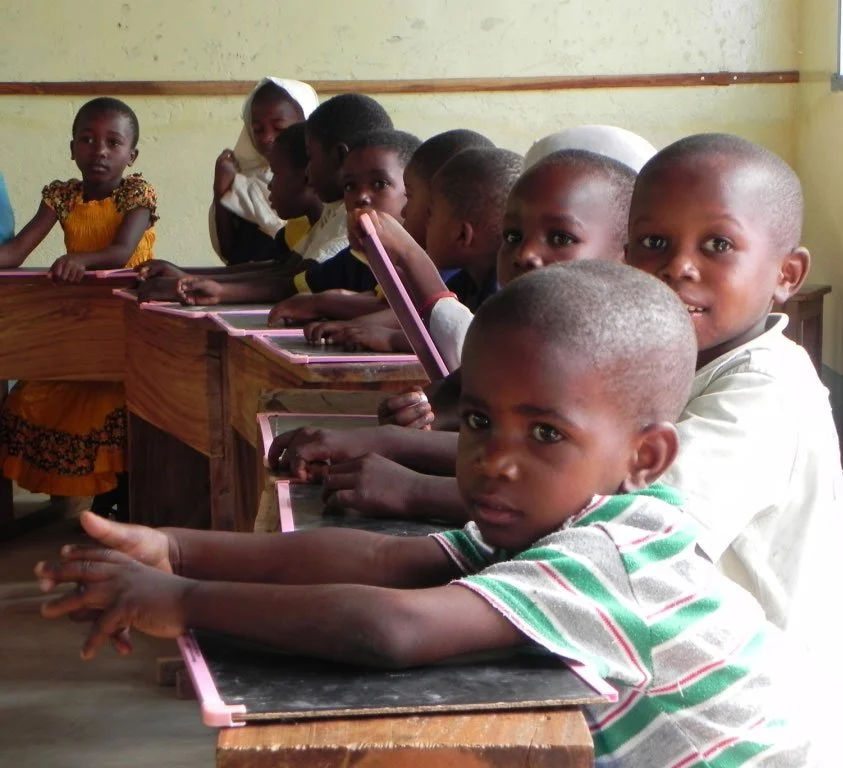 A classroom with young children sitting at wooden desks, facing forward, with some holding pink writing chalk and looking at the camera.