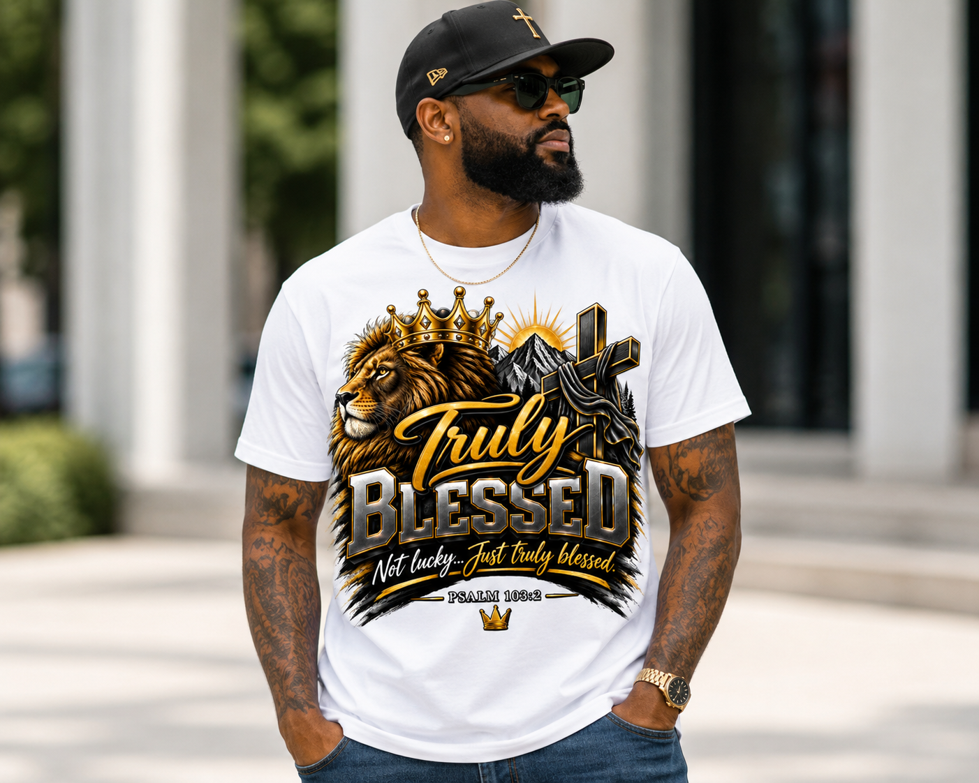 A man wearing sunglasses, a black cap with gold cross, and a graphic t-shirt stands outdoors. The t-shirt features a lion with a crown, a mountain, and the text 'Truly Blessed' with a Bible verse reference.