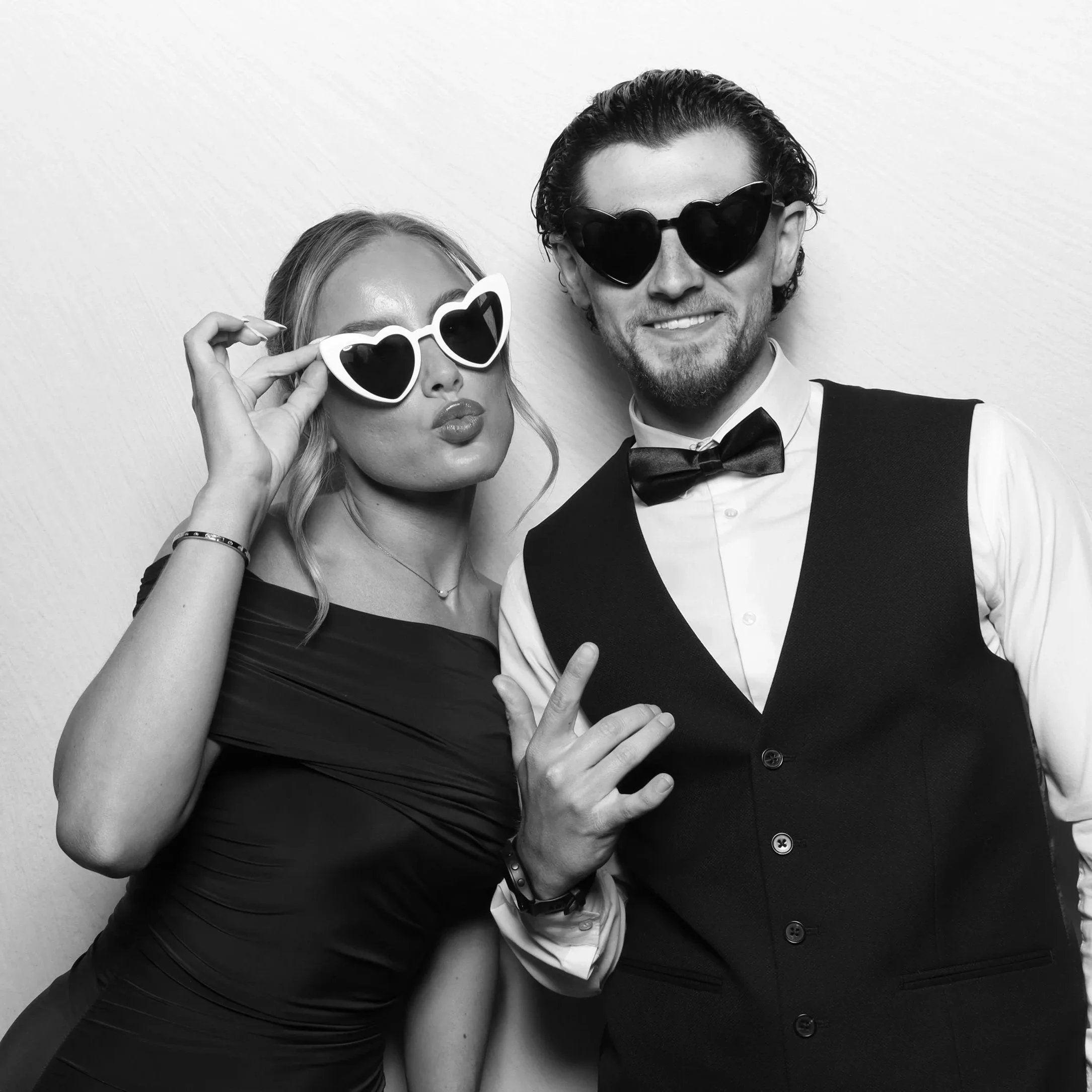 A man and woman wearing heart-shaped sunglasses and formal attire, posing against a plain background.