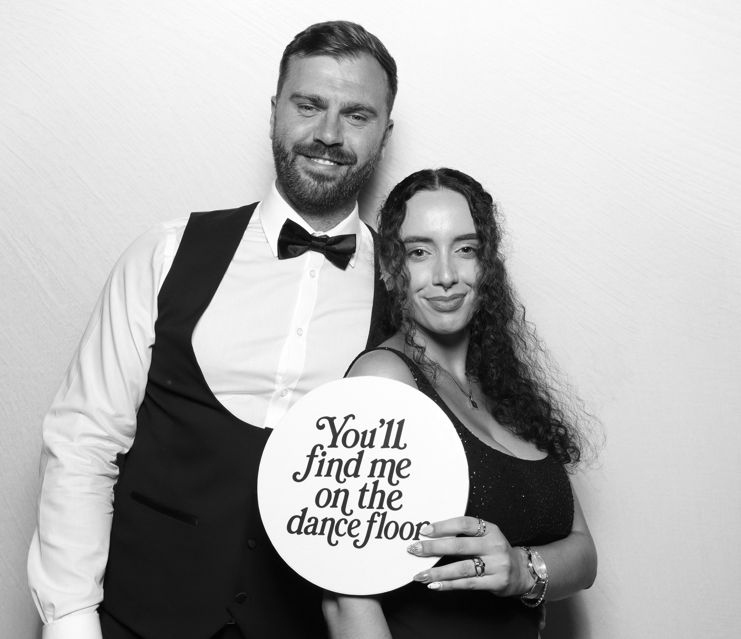 Black and white photo of a smiling man wearing a tuxedo with bow tie and a woman holding a sign that reads "You'll find me on the dance floor."
