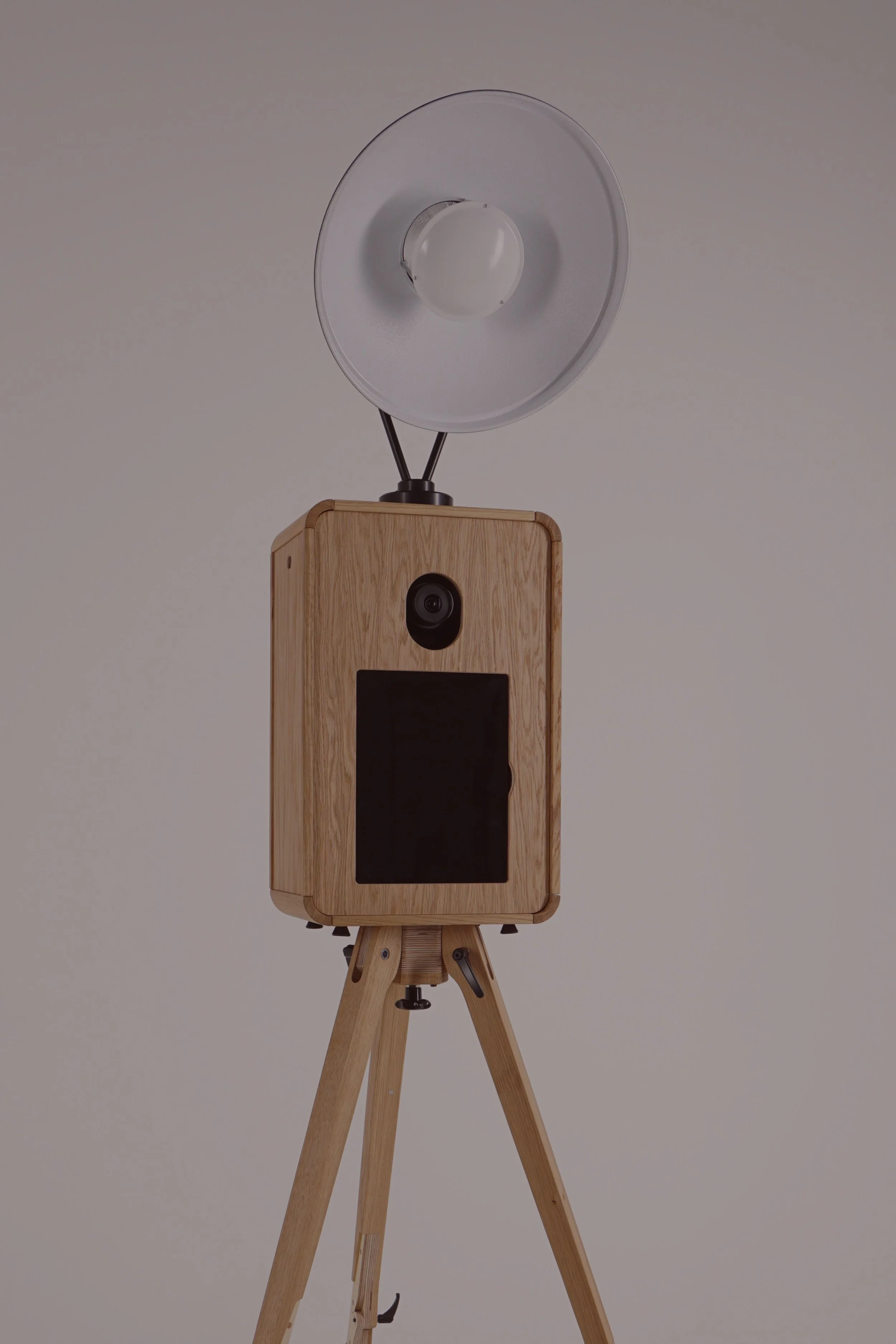 A wooden open-air photo booth with a camera and iPad feature, supported by a wooden tripod style stand, with a large circular beauty dish light fixture.